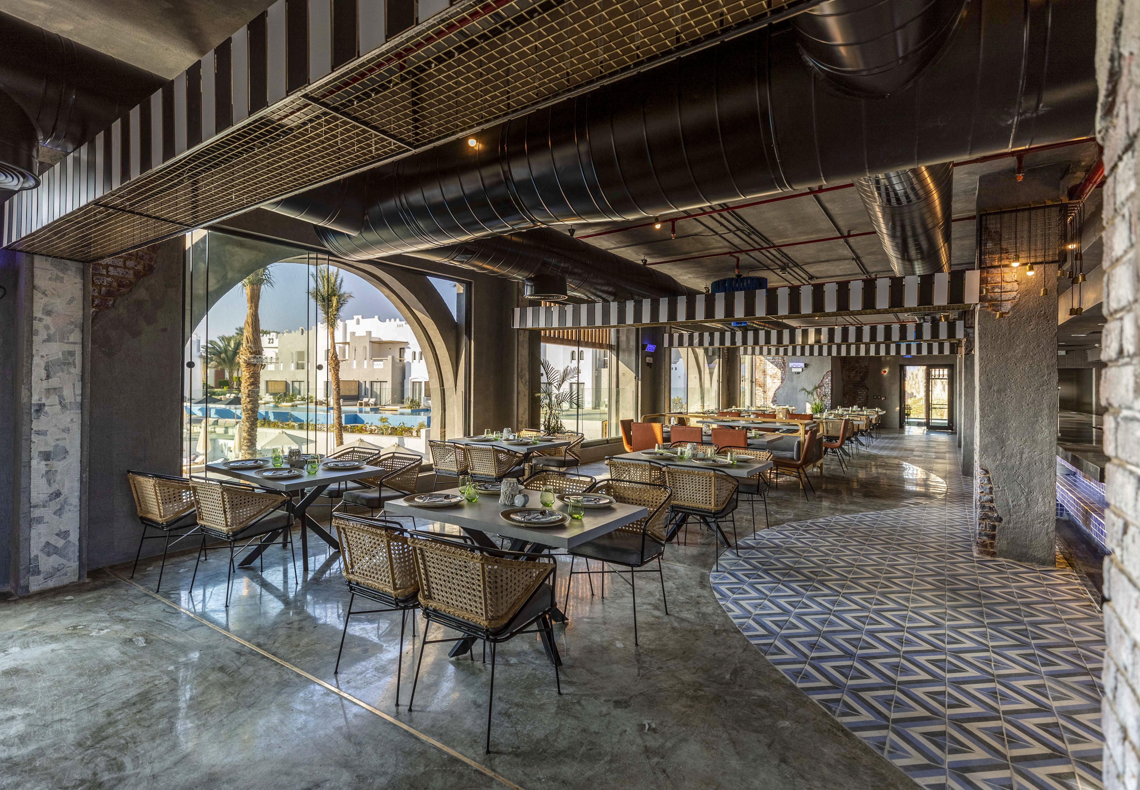 Contemporary restaurant seating is arranged around tables in an open, airy interior with large arched openings. The space overlooks an outdoor pool and courtyard with palm trees and surrounding buildings. Industrial ceiling ducts, striped accents, and geometric patterned floor tiles create a stylish, modern atmosphere. Natural light floods the area, highlighting the mix of metal and wicker furniture and polished concrete flooring.
