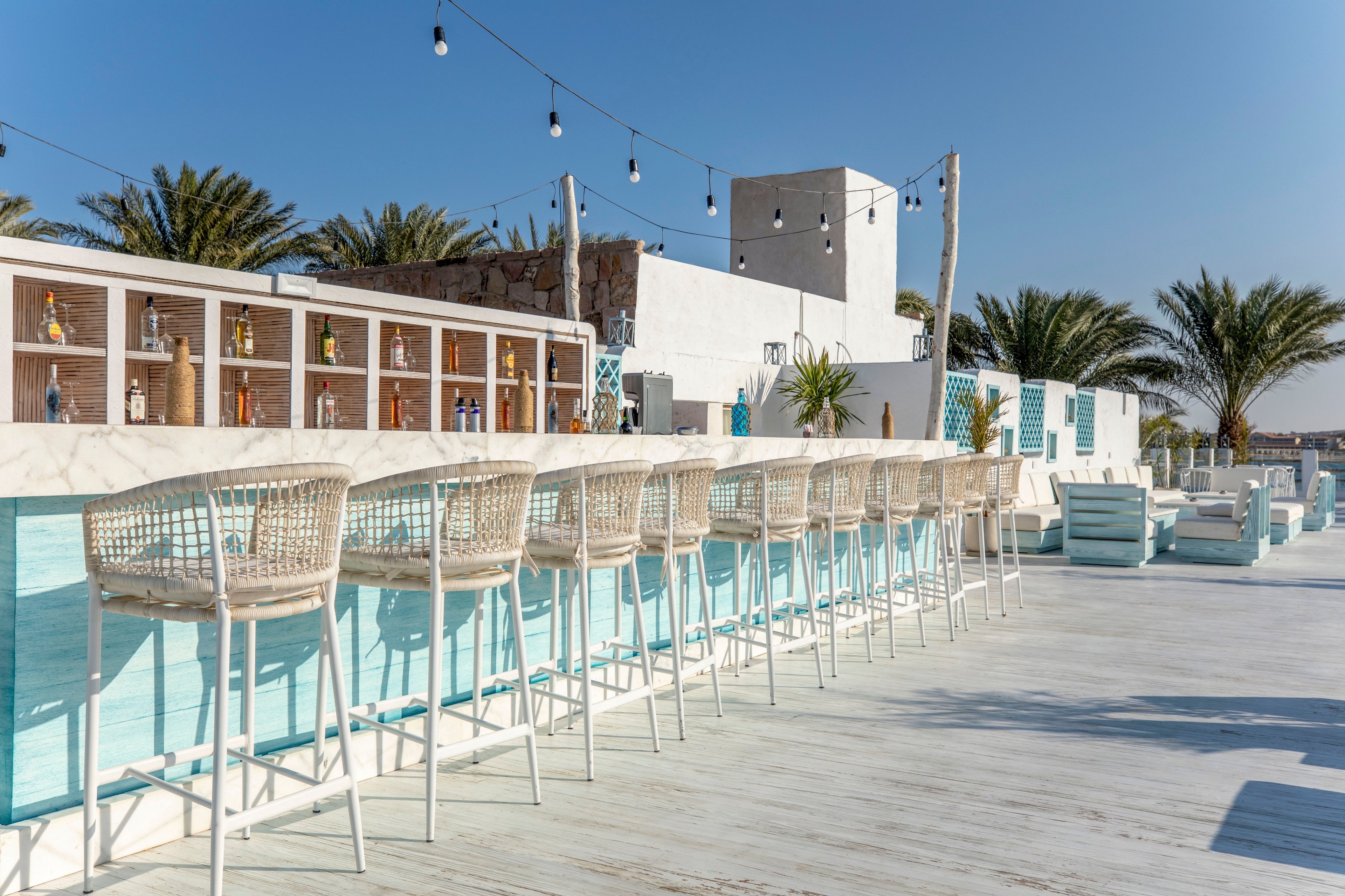 An open air bar with high wicker stools lines a long counter in a bright coastal setting. White and light blue tones dominate the modern outdoor space, complemented by palm trees in the background. Bottles are neatly arranged on shelves behind the bar, and string lights hang overhead. Lounge seating is visible further back, creating a relaxed, resort-like atmosphere under a clear blue sky.