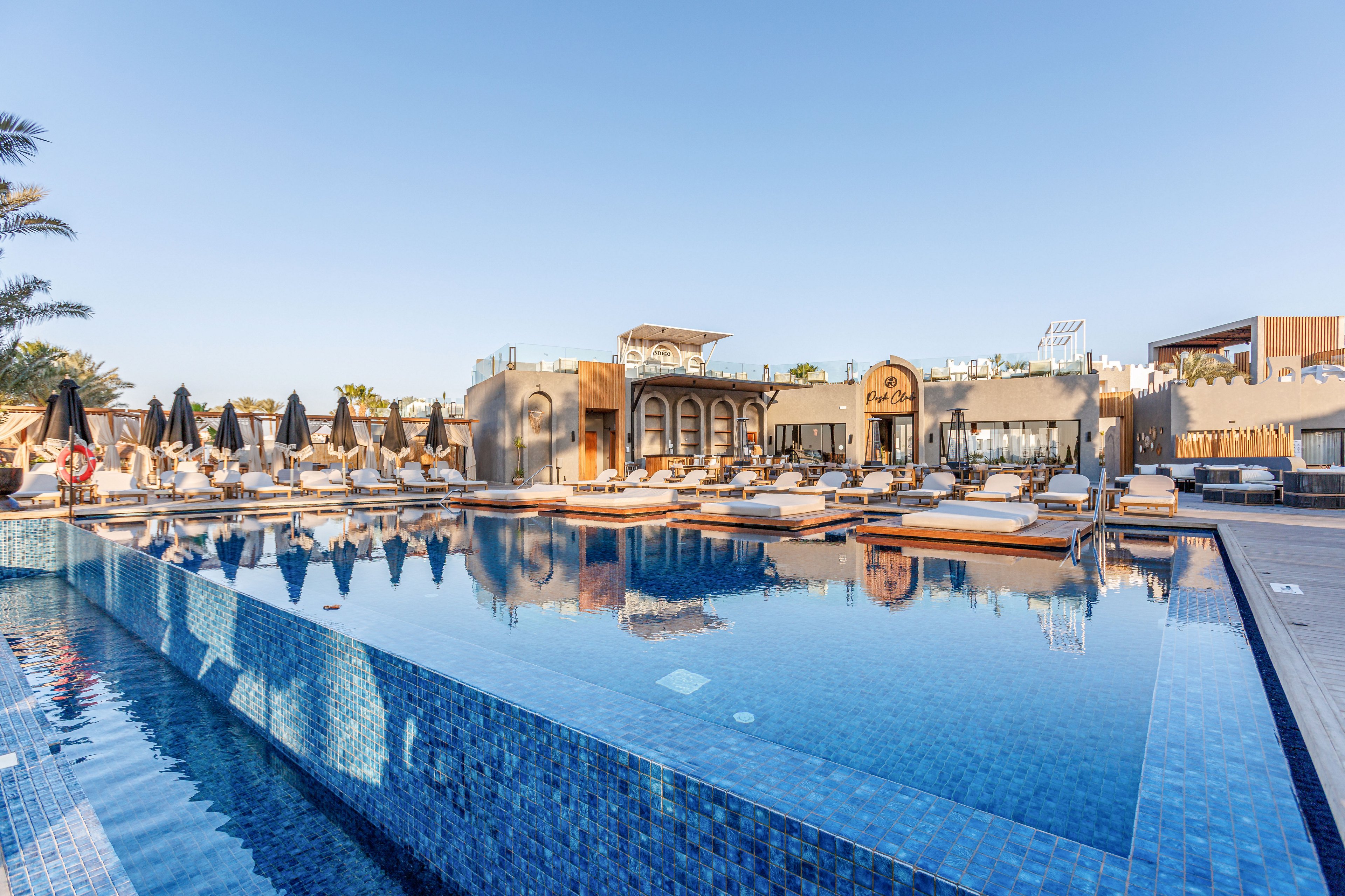 A large, clear blue swimming pool reflects a luxurious resort building under a bright morning sky. Sun loungers with cushions and umbrellas line the poolside, suggesting a relaxing vacation setting. The architecture features arches and warm-toned facades, adding a Mediterranean or Middle Eastern aesthetic. Palm trees and outdoor seating areas enhance the tranquil, upscale atmosphere.