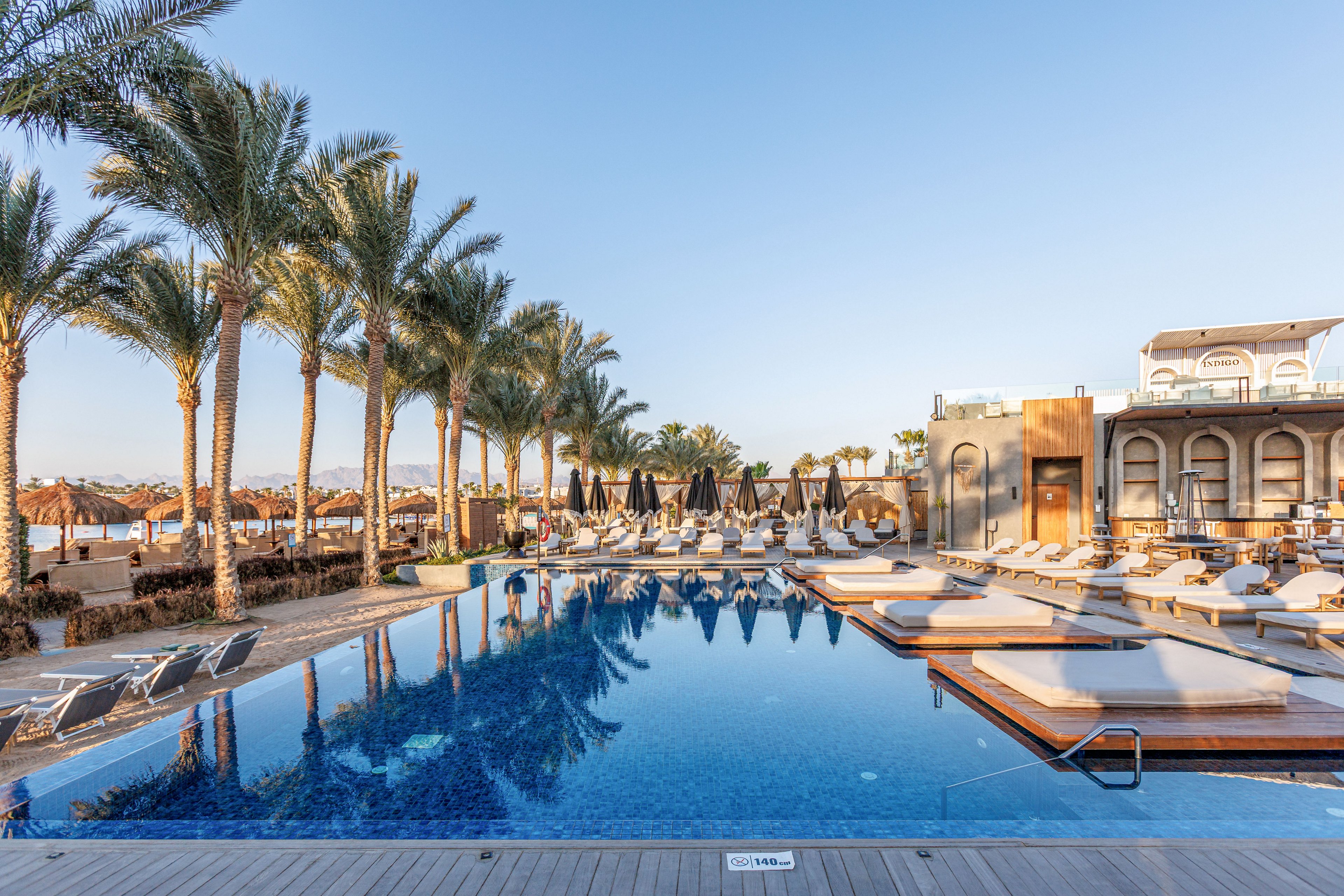 A large, tranquil swimming pool reflects tall palm trees and a clear blue sky at a luxury resort. Sun loungers and daybeds line the poolside, many shaded by umbrellas, creating a relaxing vacation atmosphere. A modern building with arched windows and warm-toned walls stands to the right, suggesting hotel accommodations or a pool bar. The scene is calm and quiet, with no visible guests, emphasizing serenity and early morning light.