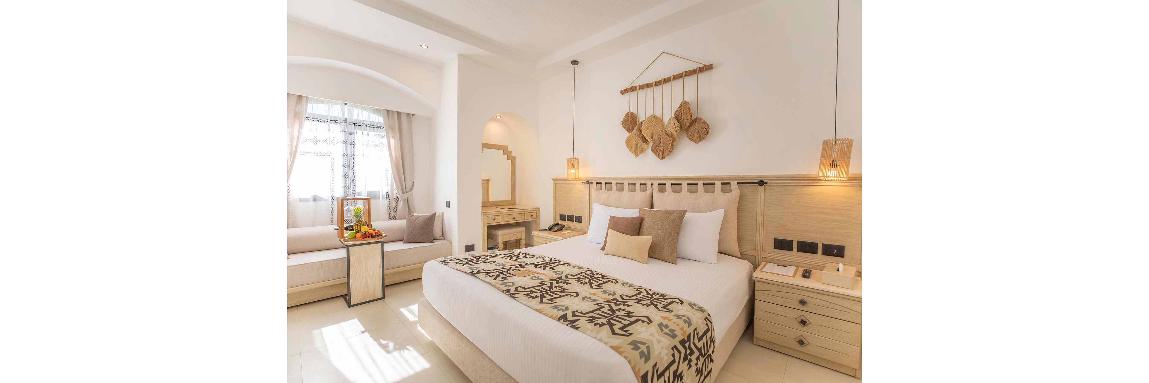 A spacious hotel bedroom features a large bed with patterned bedding and multiple accent pillows. The room is decorated in light neutral tones with wooden furniture and soft ambient lighting. A built-in sofa by the window holds a tray of fresh fruit, adding a touch of color. Natural light filters through sheer curtains, creating an airy and inviting atmosphere ideal for relaxation.