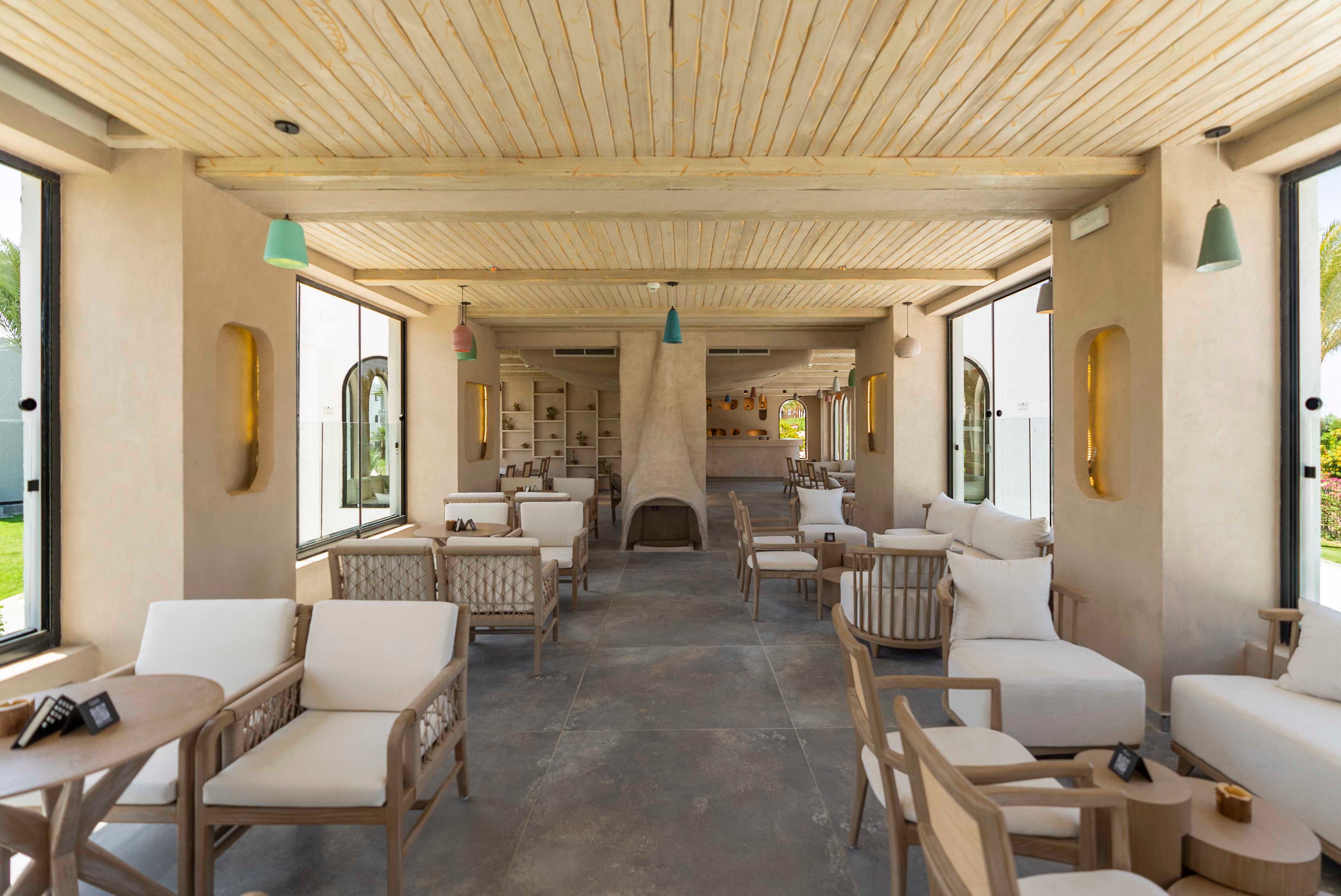 Spacious indoor lounge area featuring light wood ceilings and neutral upholstered seating. Large open windows on both sides bring in natural light and views of greenery outside. A central fireplace and bar area are visible at the far end of the room. Soft beige tones, minimalist furniture, and subtle pendant lighting create a relaxed, contemporary resort atmosphere.