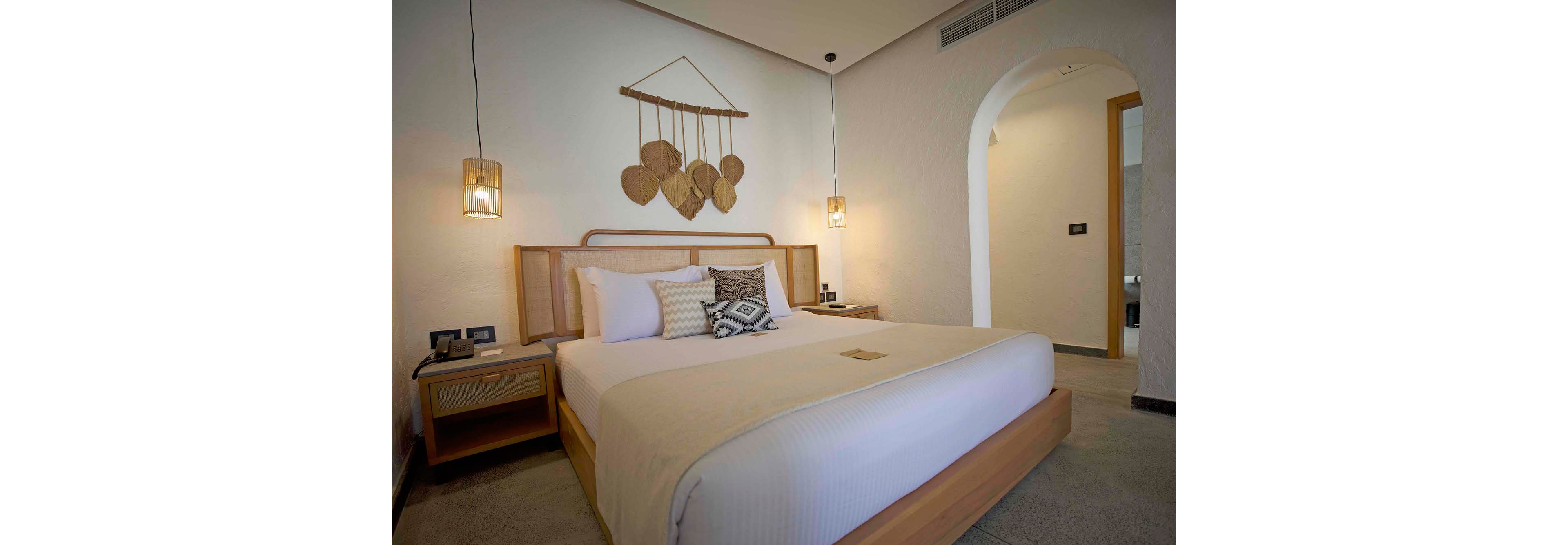 A contemporary hotel bedroom features a large neatly made bed with neutral-toned linens and patterned accent pillows. Wooden bedside tables with matching lamps flank the bed, casting a warm ambient light. A decorative wall hanging made of natural materials adds texture above the headboard. The room has a soft, minimalist aesthetic with light-colored walls, an arched doorway, and subtle modern finishes.