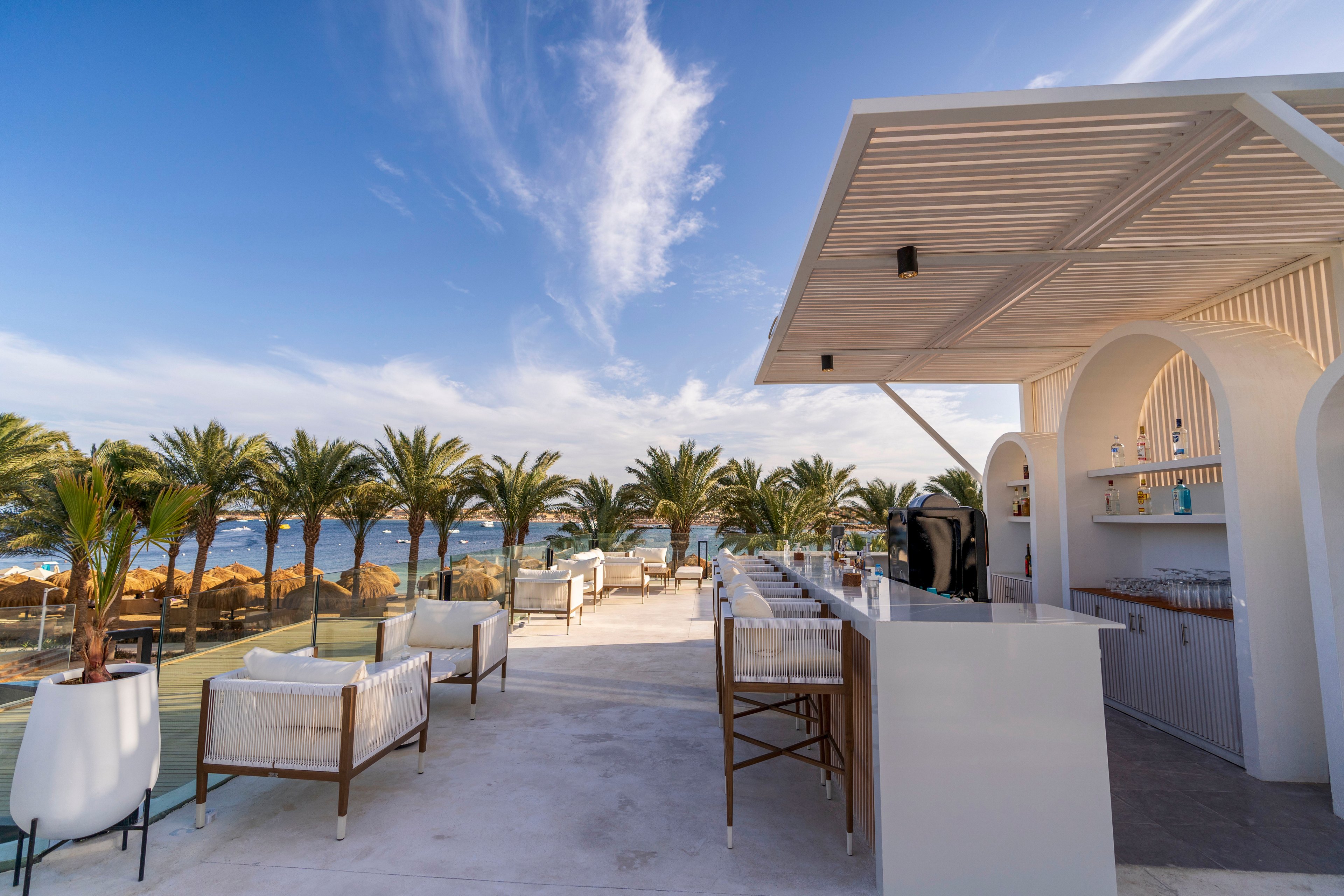 Open-air bar terrace overlooks a scenic beachfront lined with palm trees. White lounge chairs and bar stools are arranged along a sleek counter under a modern pergola. The setting is bright and sunny with clear blue sky and wispy clouds above. Overall, the scene conveys a relaxed, luxurious coastal atmosphere ideal for outdoor socializing.