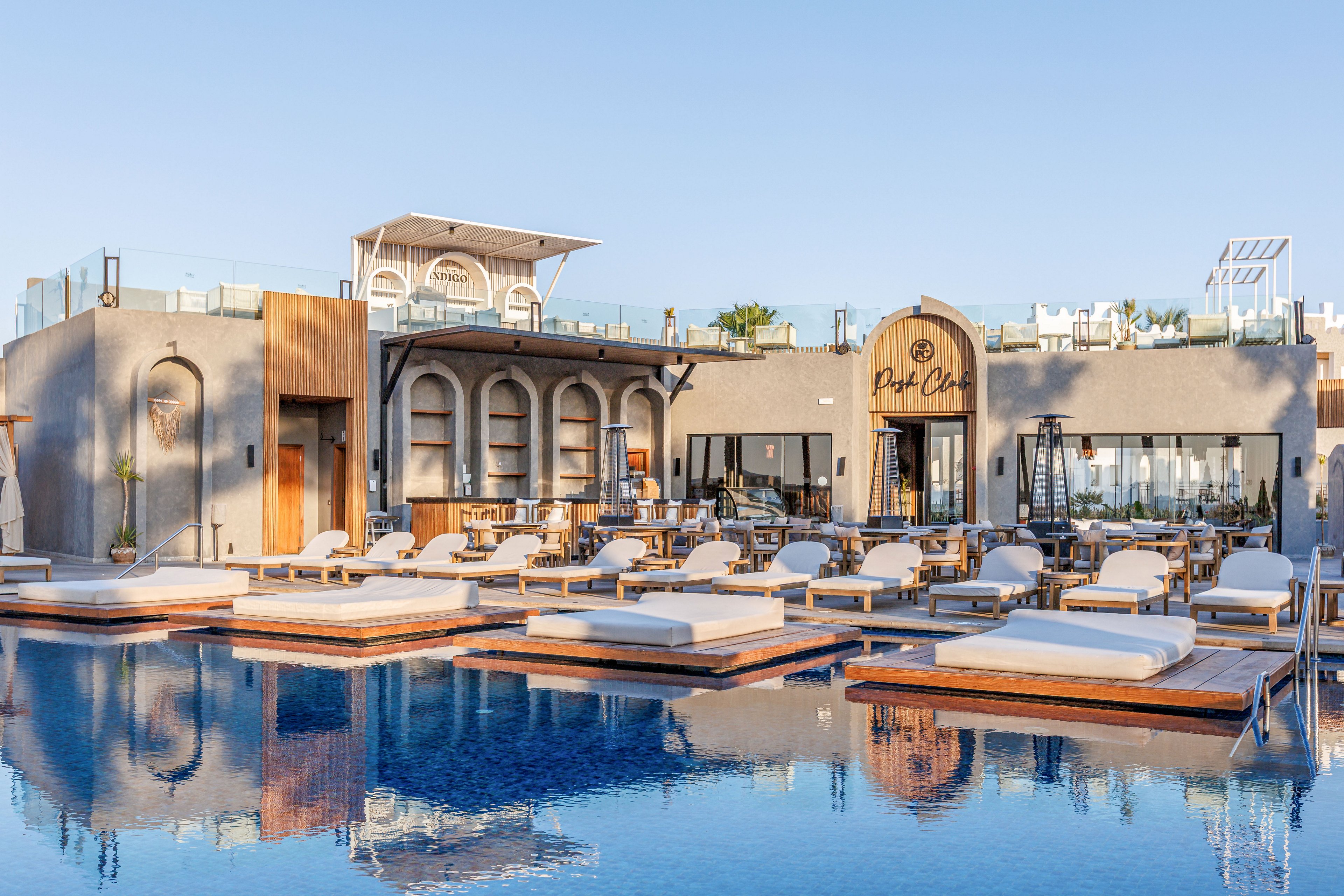A contemporary beach club features a serene swimming pool lined with cushioned sun loungers and wooden decks. The architecture combines clean lines with arched details and large glass doors, creating a stylish resort atmosphere. Warm sunlight reflects off the calm blue water, enhancing the relaxed, luxurious mood. Outdoor seating and dining areas are visible in the background, suggesting a social yet tranquil setting.