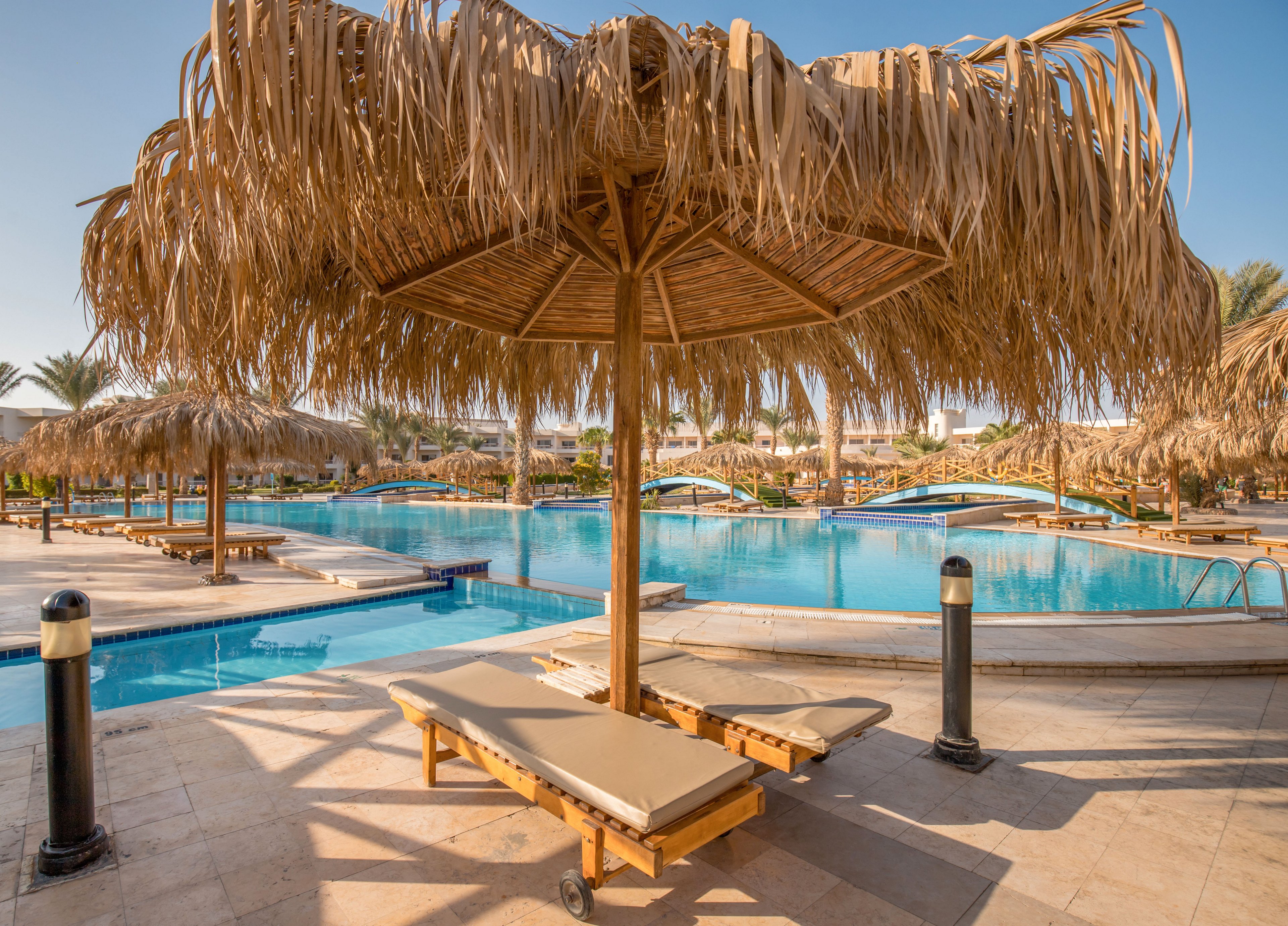 A tranquil resort swimming pool is framed by straw-covered parasols and wooden sun loungers. Clear blue water reflects the warm daylight, with small arched bridges crossing the pool in the background. Palm trees and distant buildings suggest a vacation destination in a sunny, tropical or desert climate. The scene feels calm and inviting, ideal for relaxation and leisure travel.