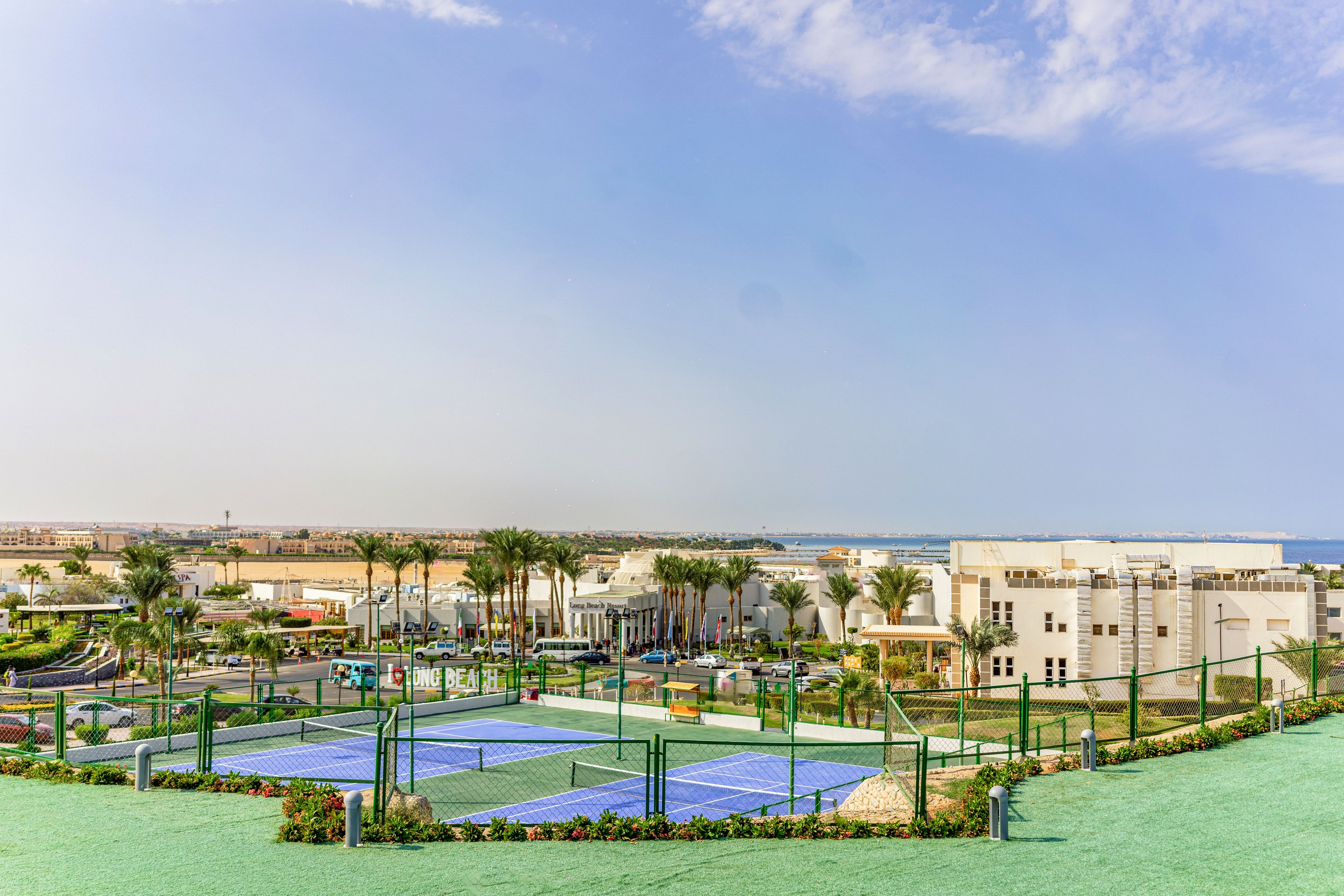 Elevated tennis courts sit in the foreground of a large seaside resort complex. Palm trees, landscaped gardens, and low-rise white buildings stretch toward the shoreline and the distant sea. The scene is bathed in bright daylight under a partly cloudy blue sky, creating a relaxed vacation atmosphere. The image conveys leisure, sports, and coastal holiday living in a modern resort setting.