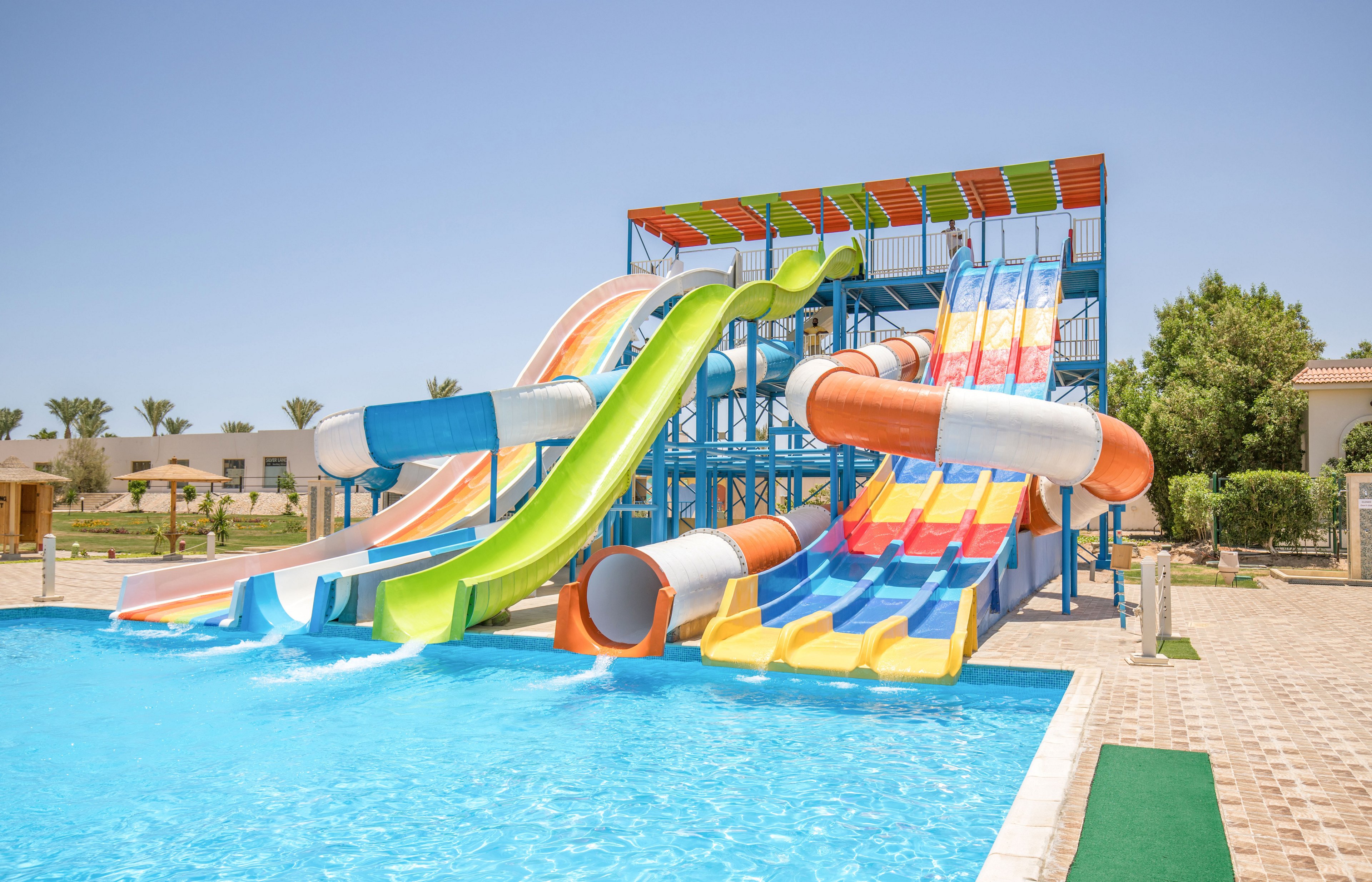 A group of large, colorful water slides descends into a bright blue swimming pool. The slides feature multiple lanes in orange, yellow, blue, green, and white, creating a playful and vibrant scene. The structure stands in an outdoor resort setting with palm trees, low buildings, and clear sunny skies in the background. The overall atmosphere suggests fun, leisure, and family-friendly recreation in warm weather.