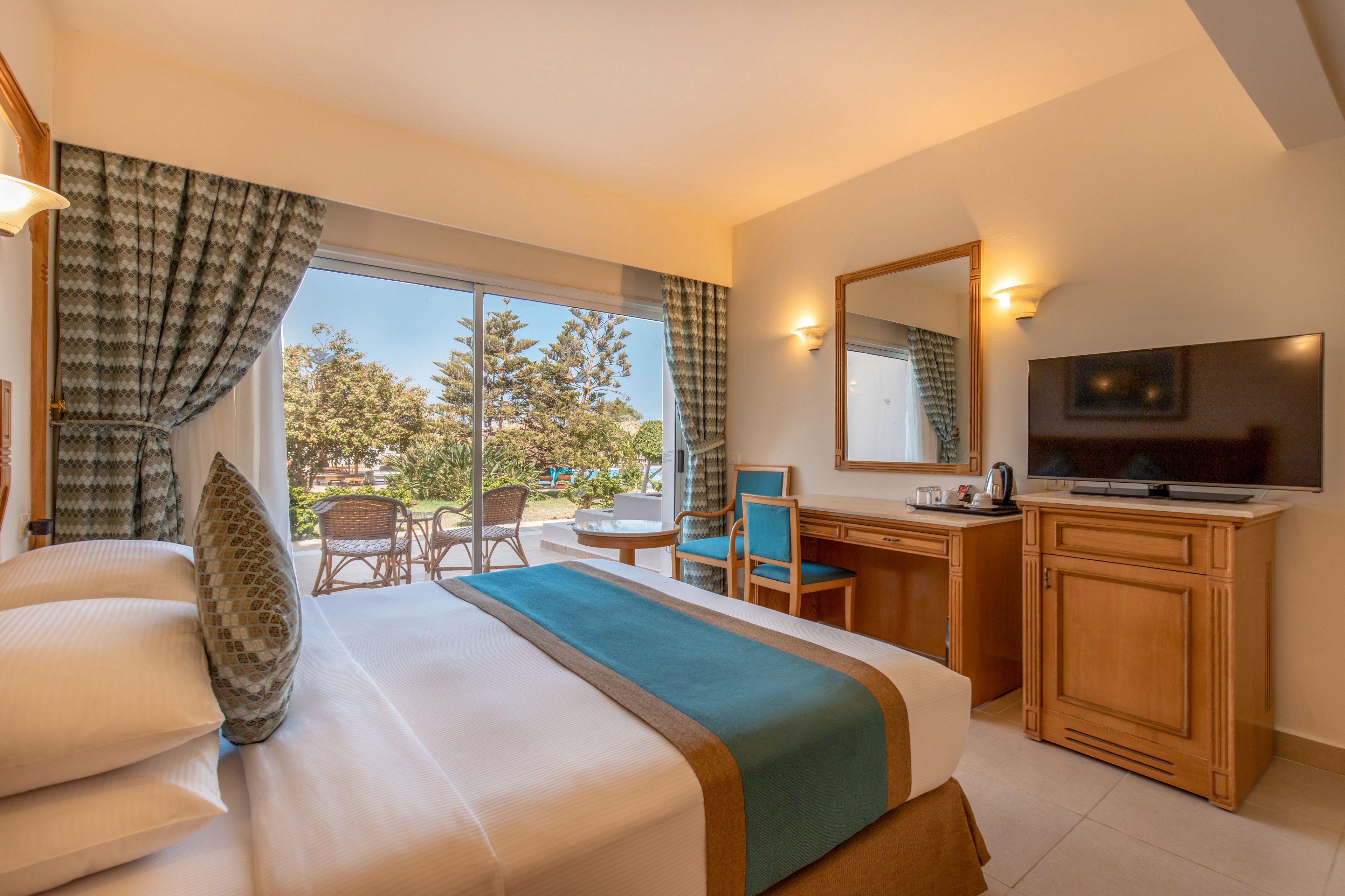 A neatly made hotel bedroom features a large bed with white linens and a teal runner. The room includes a wooden desk, mirror, flat-screen television, and a small beverage station. Floor-to-ceiling glass doors open to a balcony with outdoor chairs and a table, overlooking trees and a sunny landscape. Warm lighting and coordinated curtains create a comfortable, inviting atmosphere.