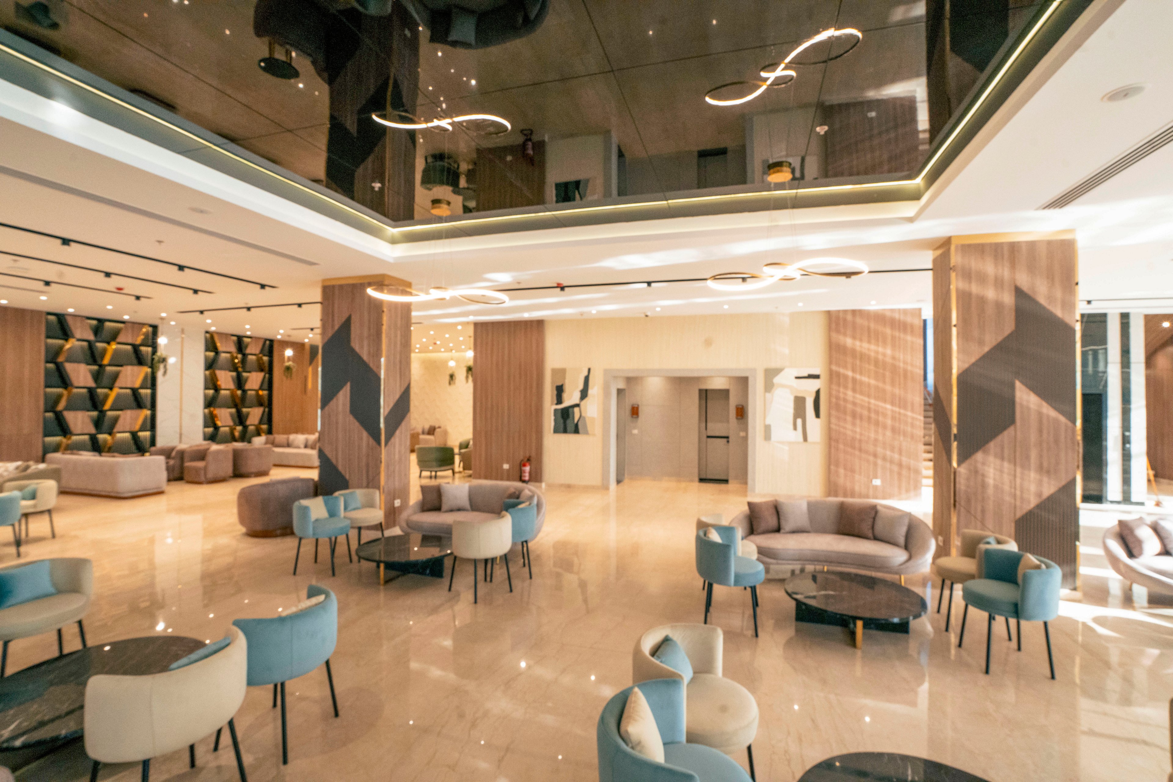 Bright contemporary lobby interior featuring curved sofas and upholstered chairs arranged around low tables. Warm wood paneling and geometric wall accents contrast with light marble flooring. A reflective ceiling with sculptural pendant lighting adds sophistication and depth to the space. The area appears open and inviting, suitable for socializing, waiting, or informal meetings.