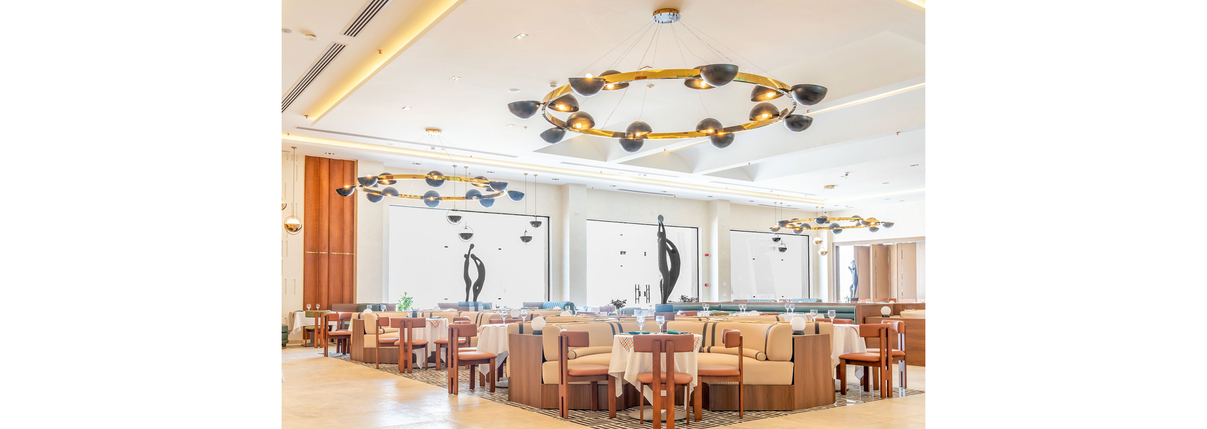 Bright contemporary restaurant dining room featuring elegant circular chandeliers and neutral-toned furniture. Tables and cushioned chairs are neatly arranged, creating an inviting and organized seating area. Large windows or wall panels display abstract black sculptures, adding artistic flair to the space. The overall atmosphere is spacious, refined, and suitable for fine dining or upscale hospitality.