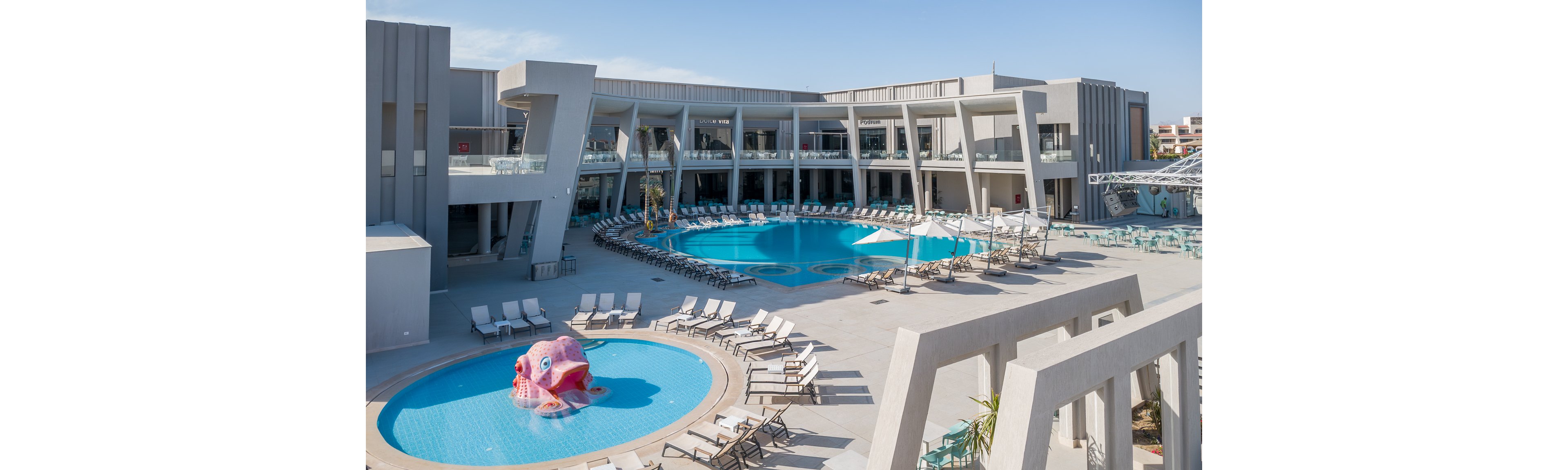 A contemporary resort complex features a large central swimming pool surrounded by a two-story building with balconies and glass railings. Numerous sun loungers and umbrellas line the pool deck, creating a relaxed vacation atmosphere. A smaller circular kids’ pool in the foreground includes a pink octopus water feature. The clear blue sky and bright daylight emphasize the clean architectural lines and leisure setting.