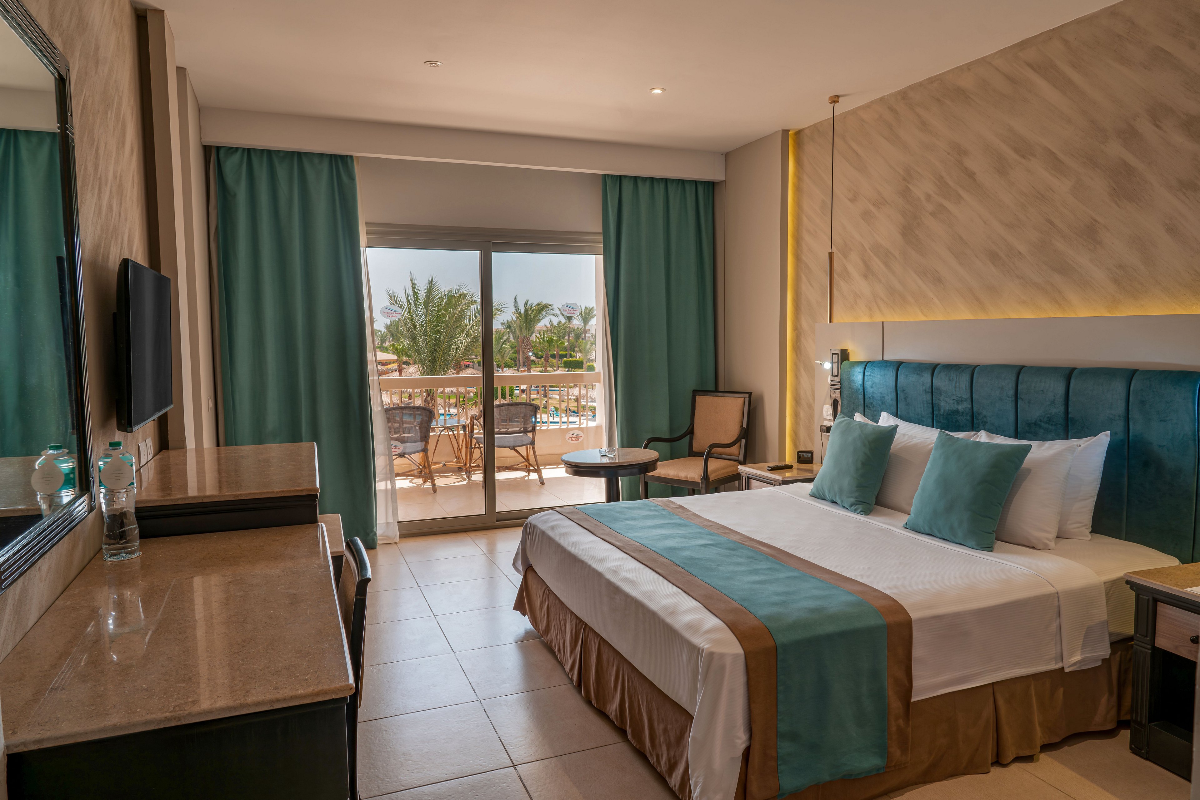 A contemporary hotel room features a large bed with teal and beige accents and matching pillows. The space includes a desk with a flat-screen TV, a chair, and a small table near the window. Floor-to-ceiling curtains frame sliding glass doors that open to a balcony overlooking a pool and palm trees. Warm lighting and neutral walls create a calm, inviting atmosphere suitable for vacation or business travel.