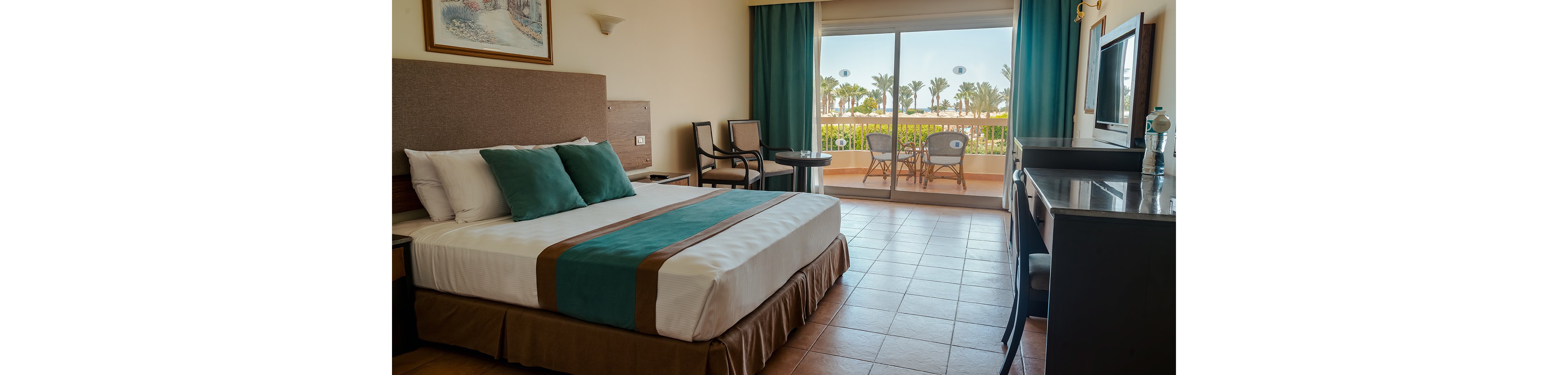 A neatly made hotel room features a large bed with teal and brown accents and matching pillows. The space includes a desk with a television, chairs, and a small table, all arranged on tiled flooring. Large sliding glass doors open to a balcony with outdoor seating and a view of palm trees and a bright, sunny sky. The overall atmosphere is clean, inviting, and ideal for a relaxing vacation stay.