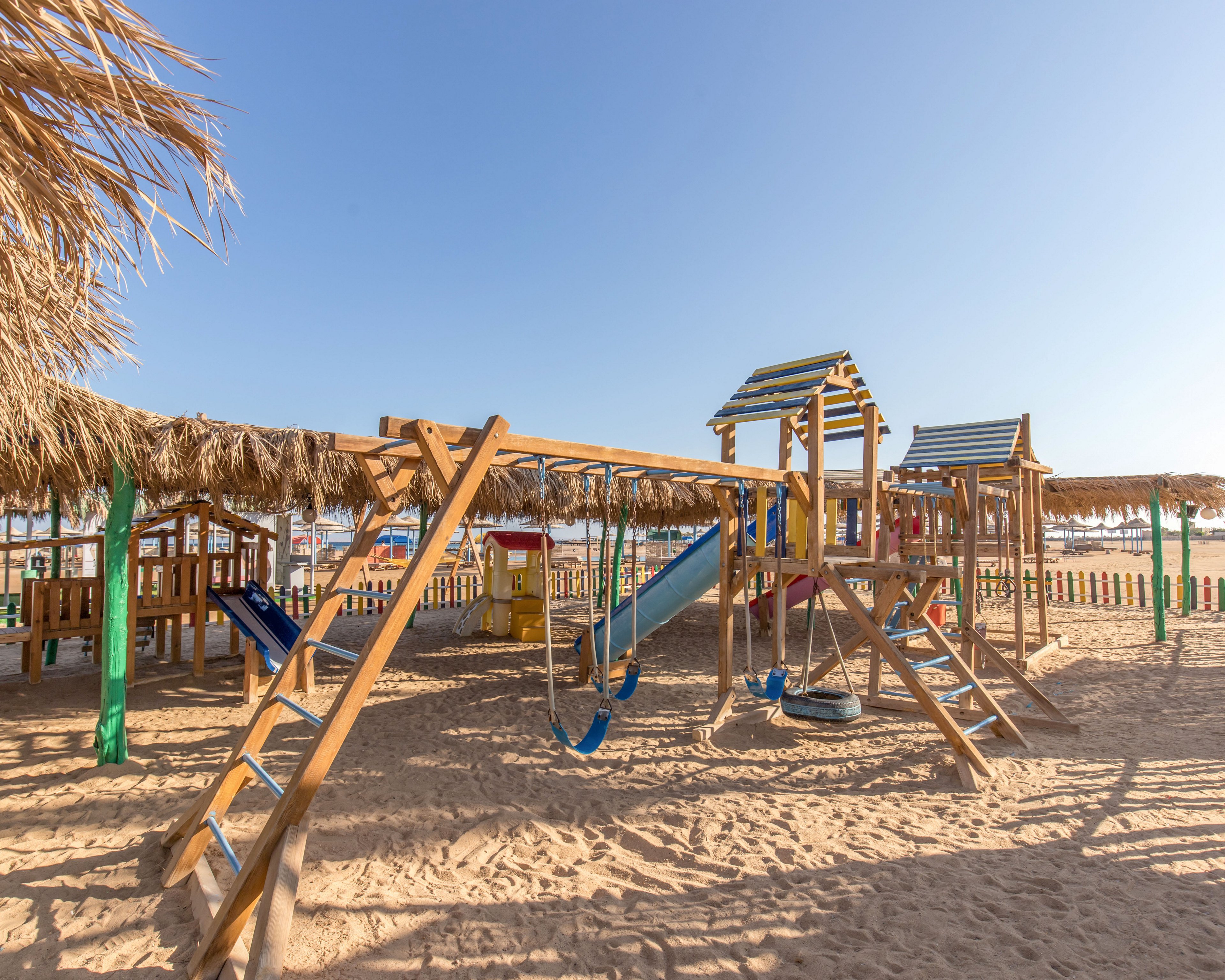 A wooden playground stands on a sandy beach under a clear blue sky. The play area includes swings, slides, climbing frames, and small huts with thatched roofs. Colorful posts and fences surround the structures, adding bright accents to the neutral sand and wood tones. The scene is empty of people, emphasizing a calm, sunny, and inviting seaside atmosphere.
