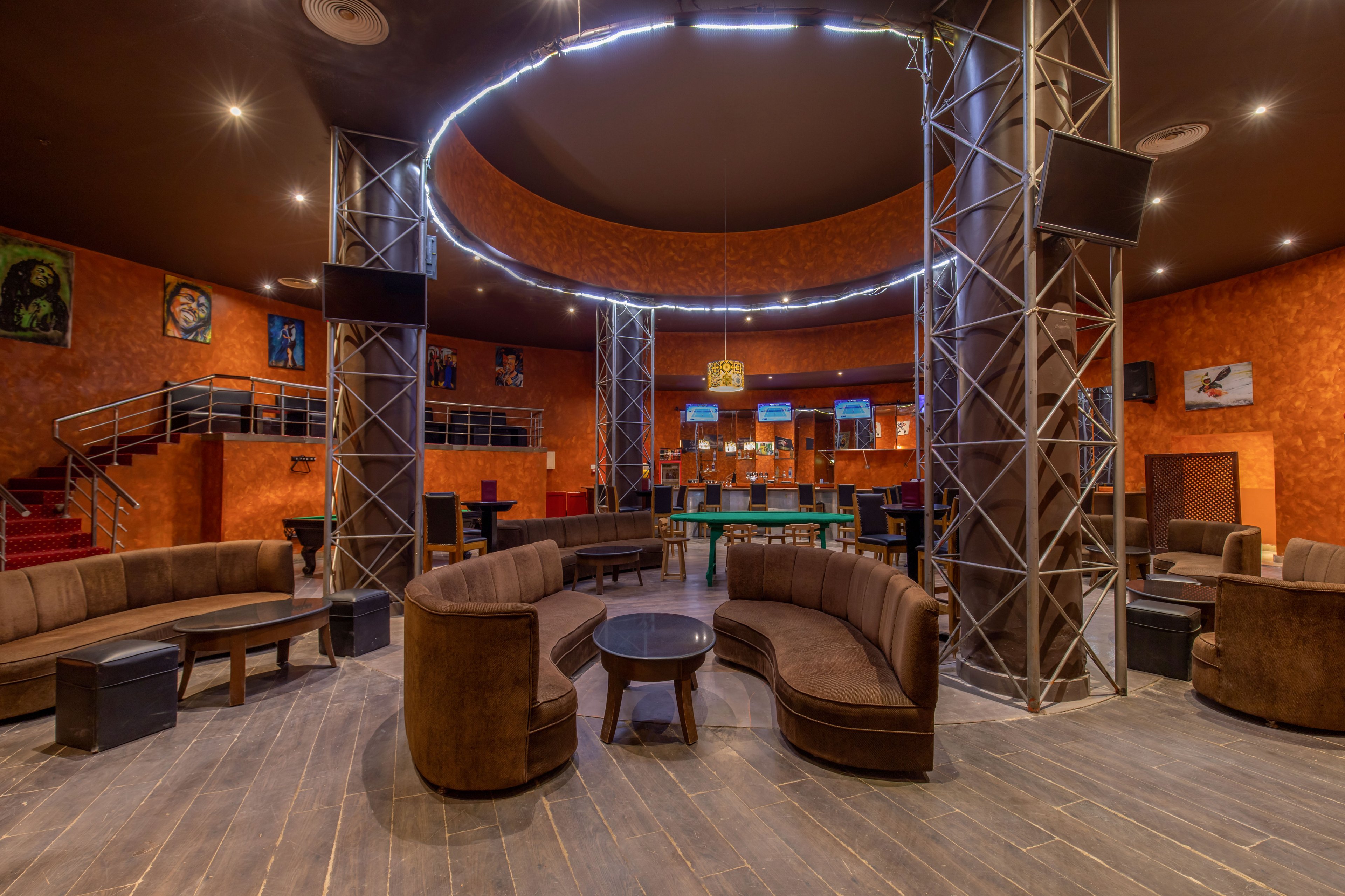 Spacious lounge bar interior featuring curved brown sofas arranged around a central circular area. Metal support columns rise to a ring of ceiling lights, creating a focal point in the room. The walls are painted a warm orange tone and decorated with framed artwork, while a bar counter with stools is visible in the background. Multiple wall-mounted screens and a mezzanine with stairs add to the contemporary entertainment atmosphere.