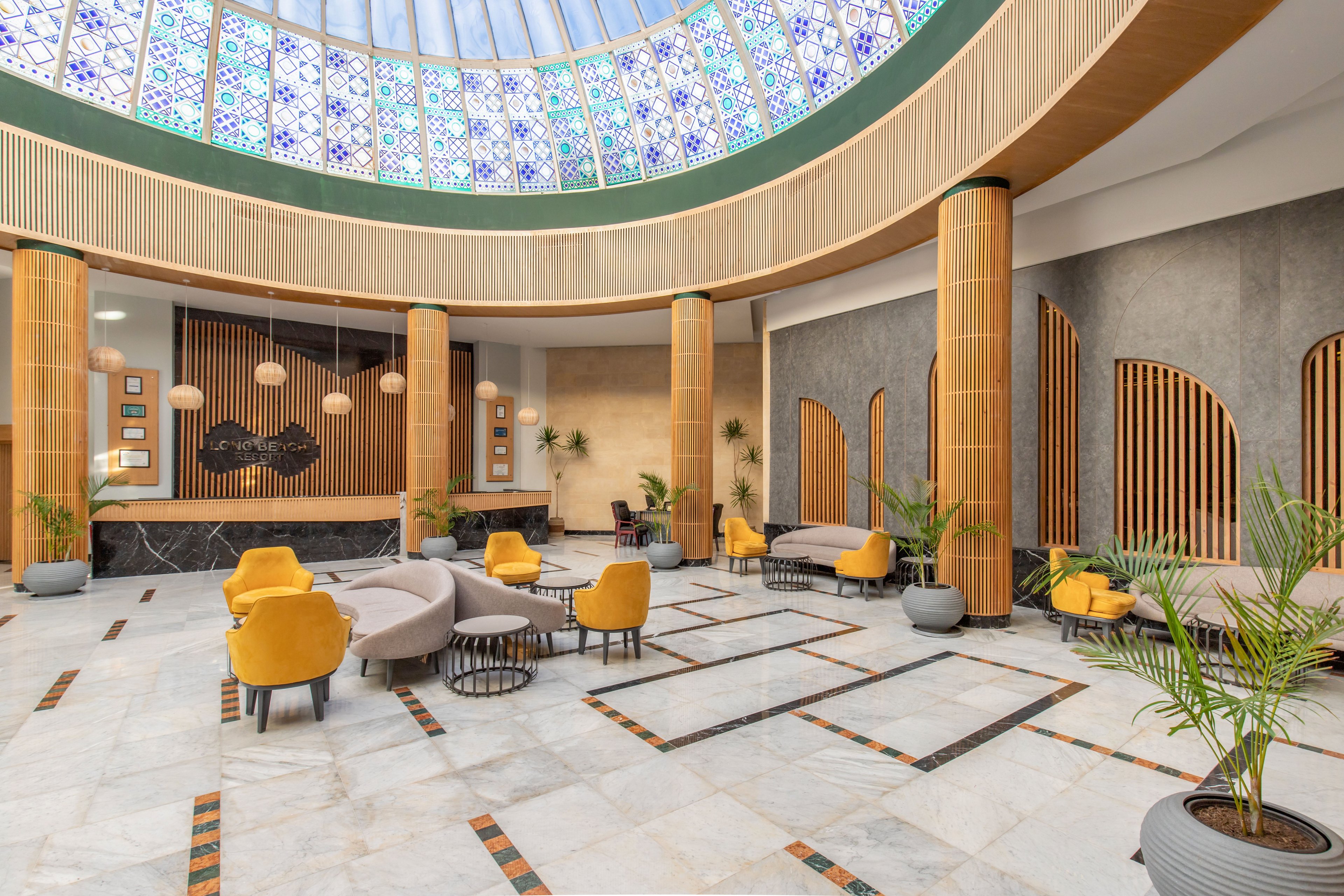 Spacious contemporary lobby interior featuring curved sofas and bright yellow lounge chairs. Tall wooden columns and decorative wall slats frame the seating area, complemented by potted plants. A large circular skylight with patterned glass illuminates the marble floor and geometric tile inlays. Hanging pendant lights and framed wall art add warmth and visual interest to the modern space.