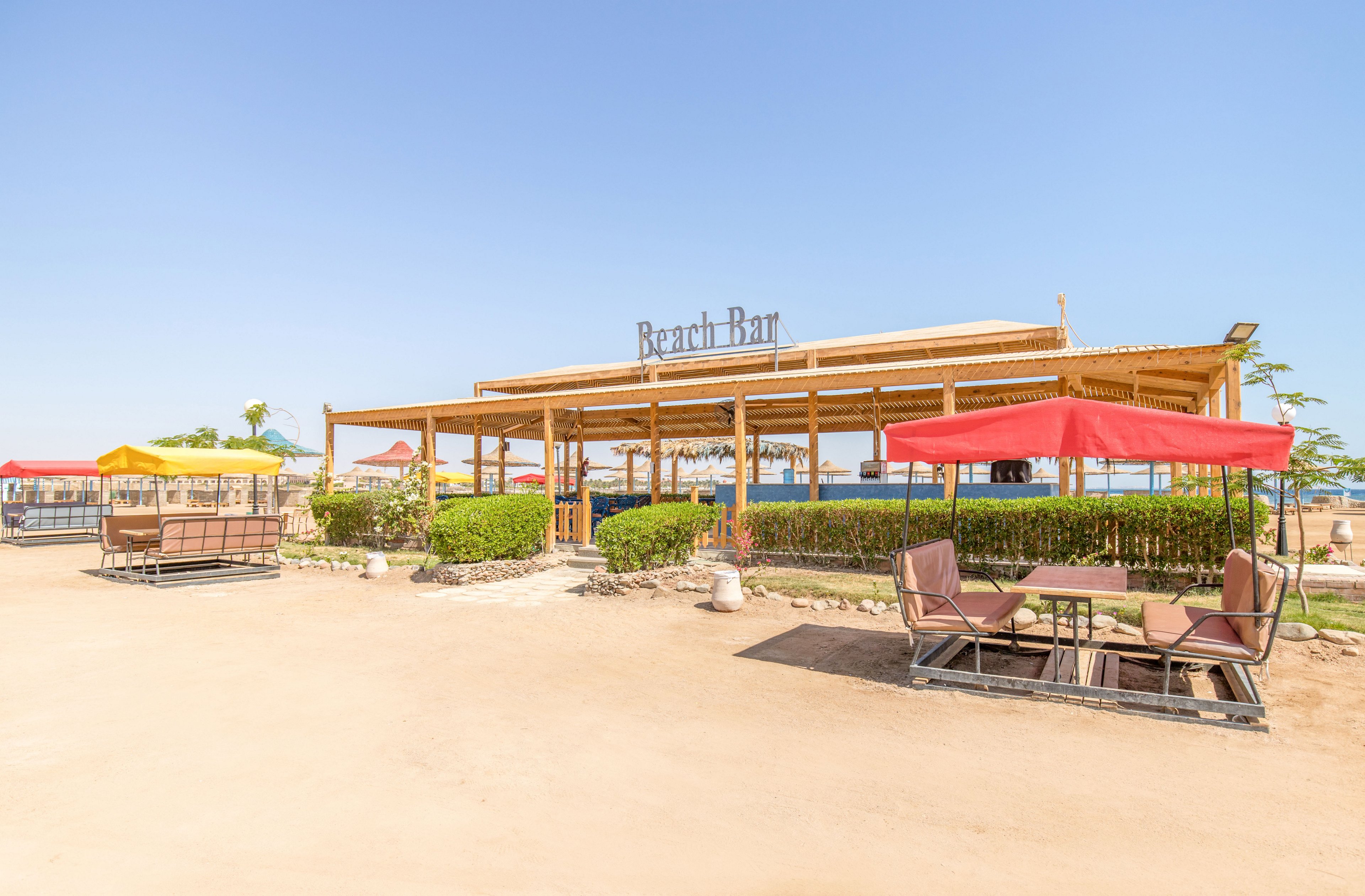 An open-air beach bar with a wooden structure stands on a sandy shoreline under a clear blue sky. The bar area is surrounded by green shrubs and shaded seating areas with colorful umbrellas. In the foreground, tables and cushioned chairs sit on a paved section, inviting guests to relax. The distant view of the sea enhances the relaxed, vacation atmosphere of the scene.