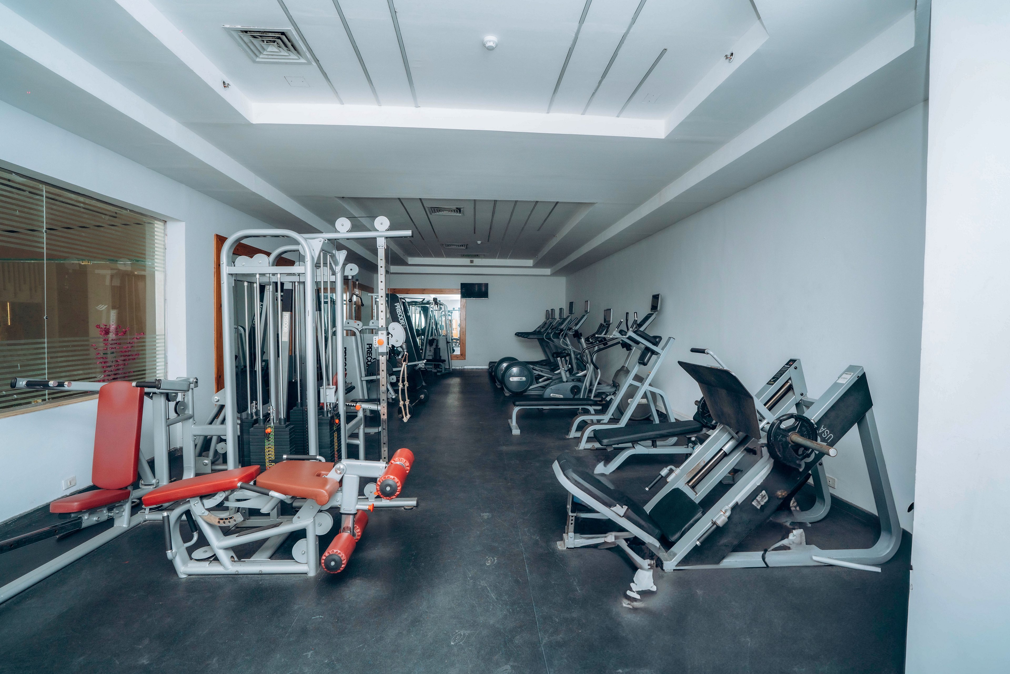 A clean, modern gym interior features various strength training machines arranged along the walls. The room includes weight benches, cable machines, and leg press equipment on a dark floor. Bright white walls and ceiling panels create a spacious, minimalist atmosphere. No people are present, emphasizing the equipment and workout environment.