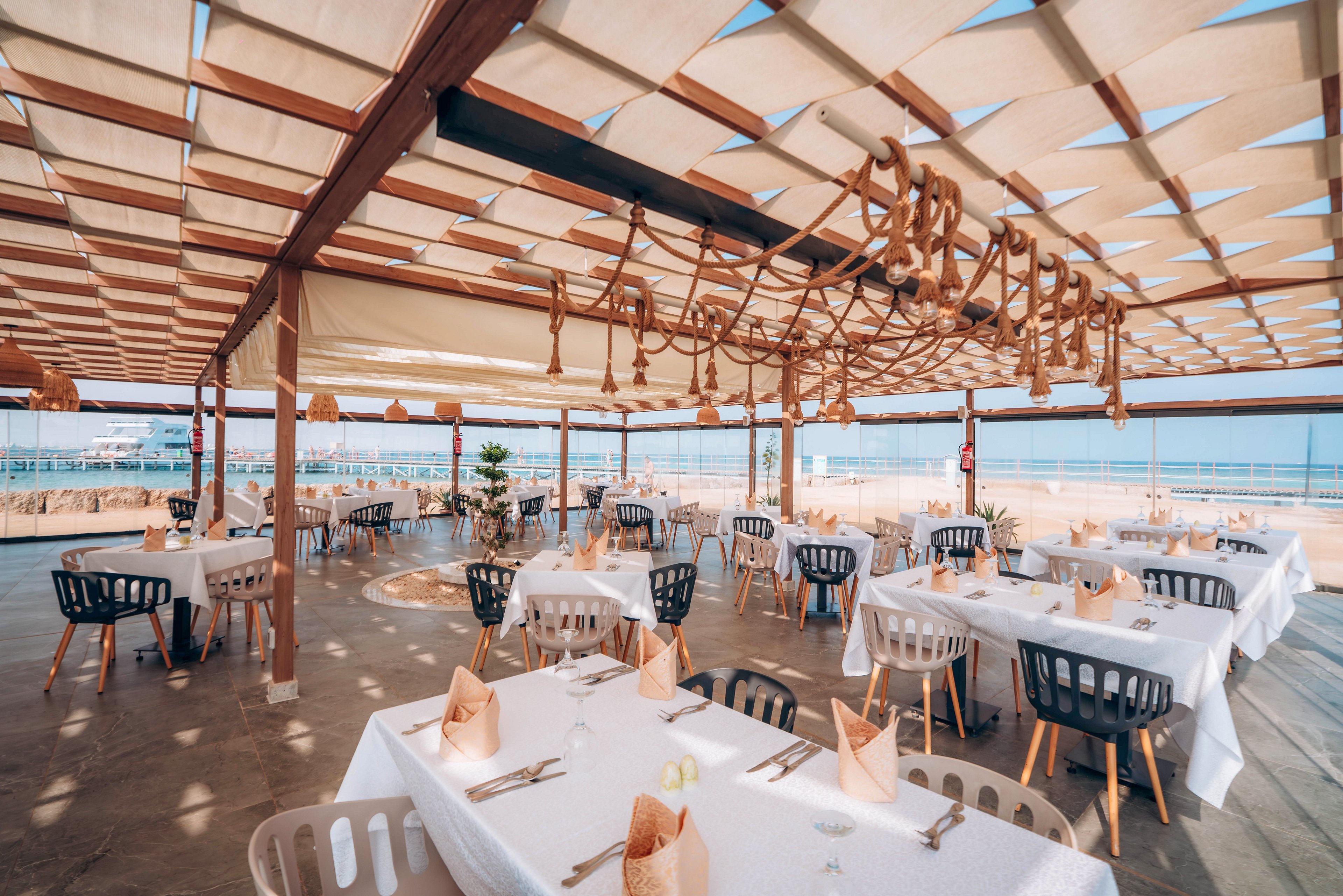 An open-air seaside restaurant is set with neatly arranged tables covered in white tablecloths. Each table features folded napkins and simple place settings, creating a clean and elegant dining atmosphere. A wooden pergola with fabric panels shades the space, while decorative rope elements hang from the ceiling. The background reveals a calm blue sea and clear sky, enhancing the relaxed coastal ambiance.