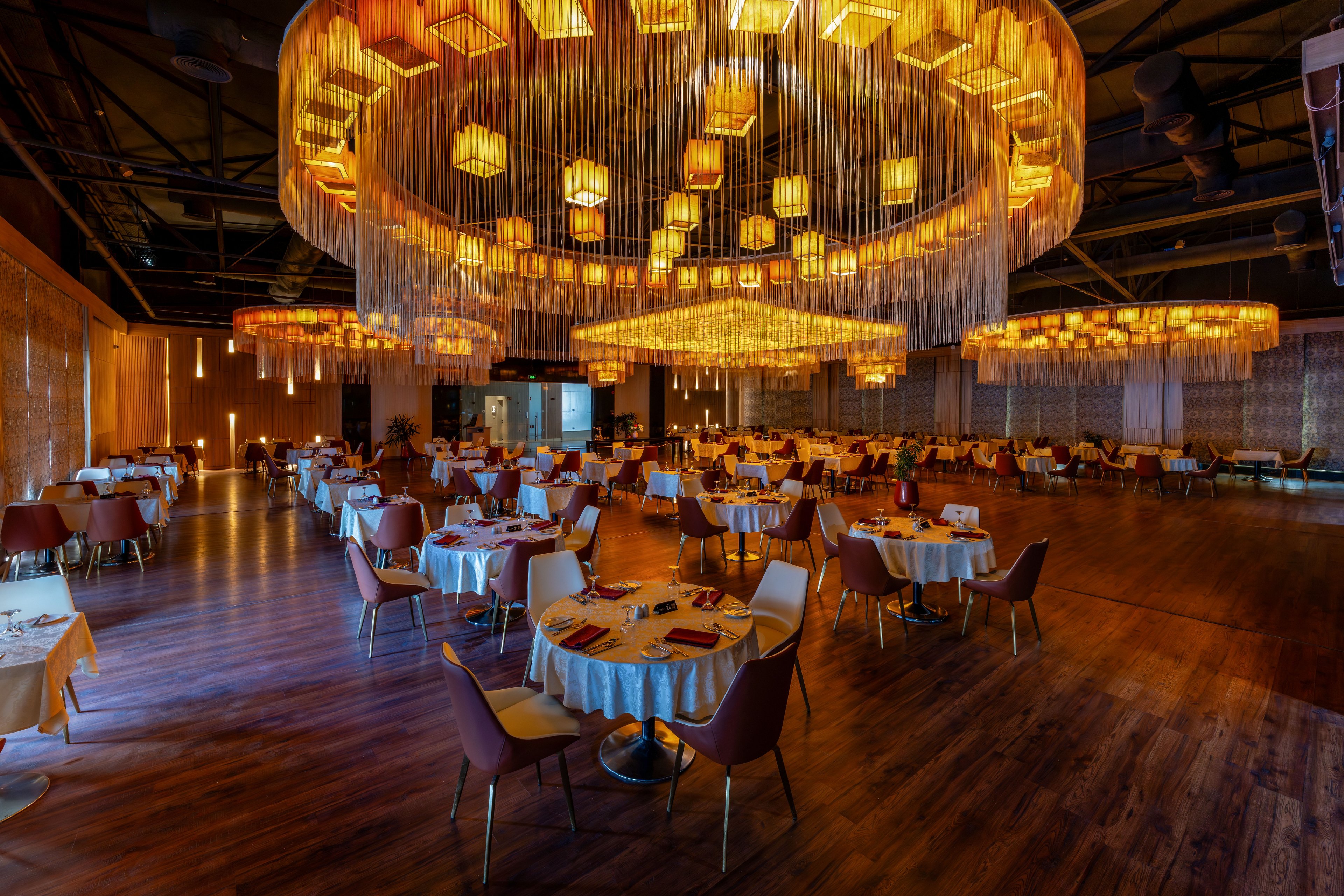 Spacious upscale restaurant dining room with round tables and modern chairs arranged across a polished wooden floor. A large circular chandelier installation with cascading strands and warm amber lights dominates the ceiling, creating a dramatic focal point. Soft golden illumination and decorative wall panels give the space a cozy, luxurious atmosphere. The room appears prepared for guests, with neatly set tables and an inviting, intimate ambiance.