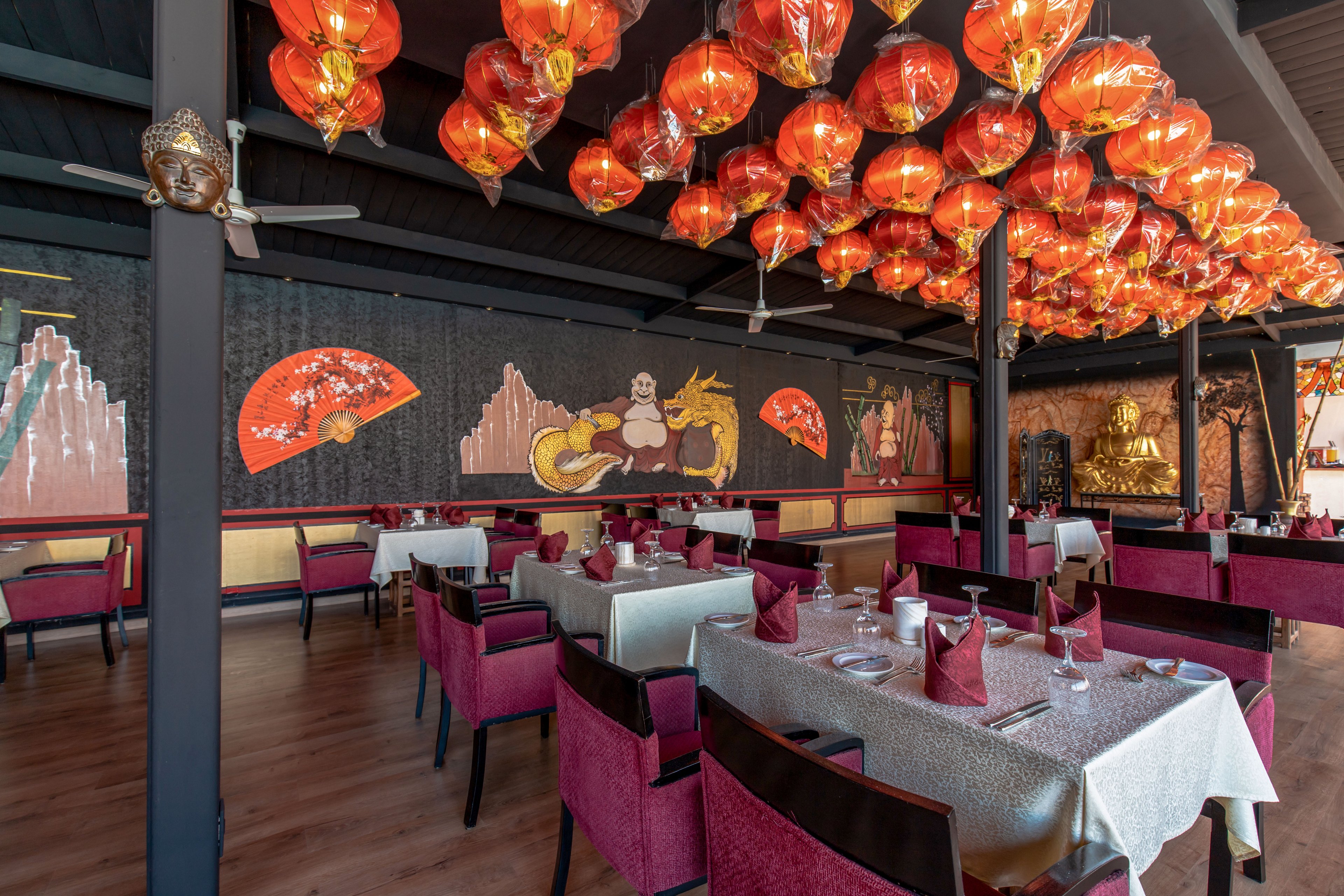 Interior of an Asian-themed restaurant featuring rows of red hanging lanterns across the ceiling. Tables are neatly set with white cloths, folded red napkins, glassware, and cutlery, creating a formal dining atmosphere. The walls display large decorative murals with fans, mountains, and a reclining figure alongside a dragon, adding cultural flair. Warm lighting, wooden flooring, and upholstered chairs in red and pink tones contribute to a cozy yet sophisticated setting.