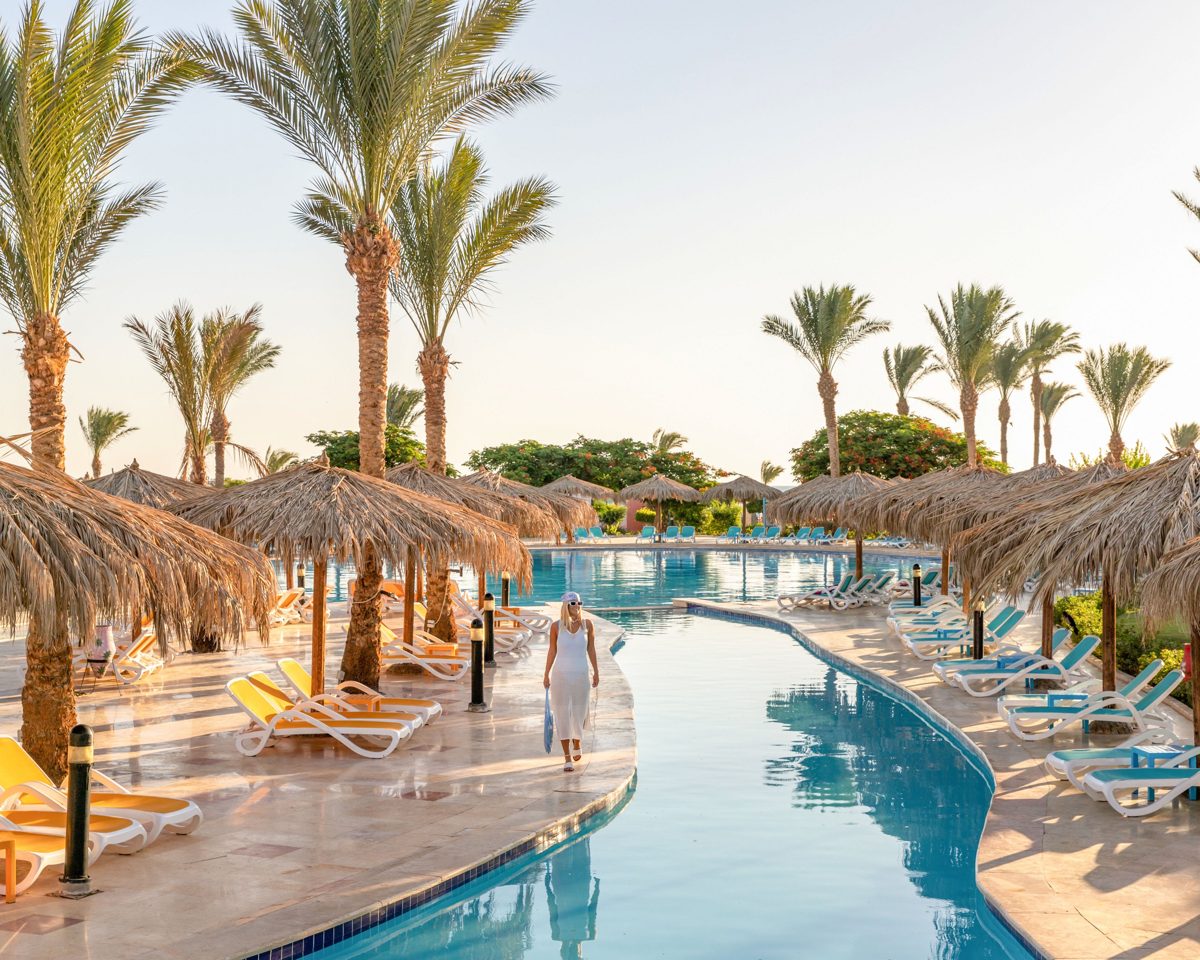 A woman in a white dress walks along a curved swimming pool at a sunny resort. Tall palm trees and thatched umbrellas line the poolside, creating a tropical atmosphere. Colorful lounge chairs are arranged neatly around the water, suggesting a relaxing vacation setting. The clear sky and warm light enhance the tranquil, holiday mood.