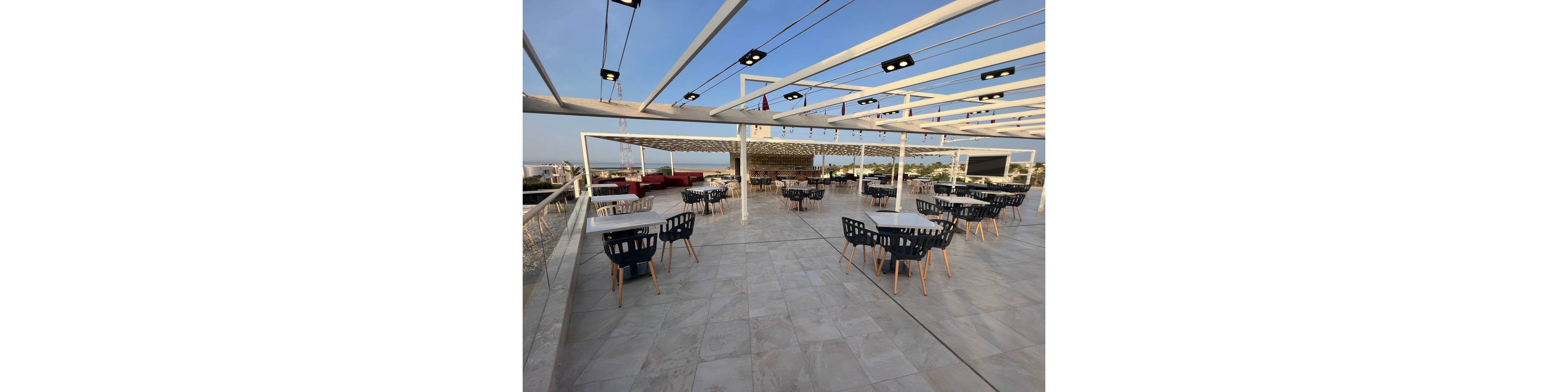 Spacious rooftop terrace features multiple tables and contemporary chairs arranged across a tiled floor. A white pergola structure with integrated lights frames the open-air dining area. Red lounge seating and additional tables appear in the background, overlooking a distant cityscape and coastline. The scene is captured in warm daylight, suggesting a relaxed, modern hospitality setting.