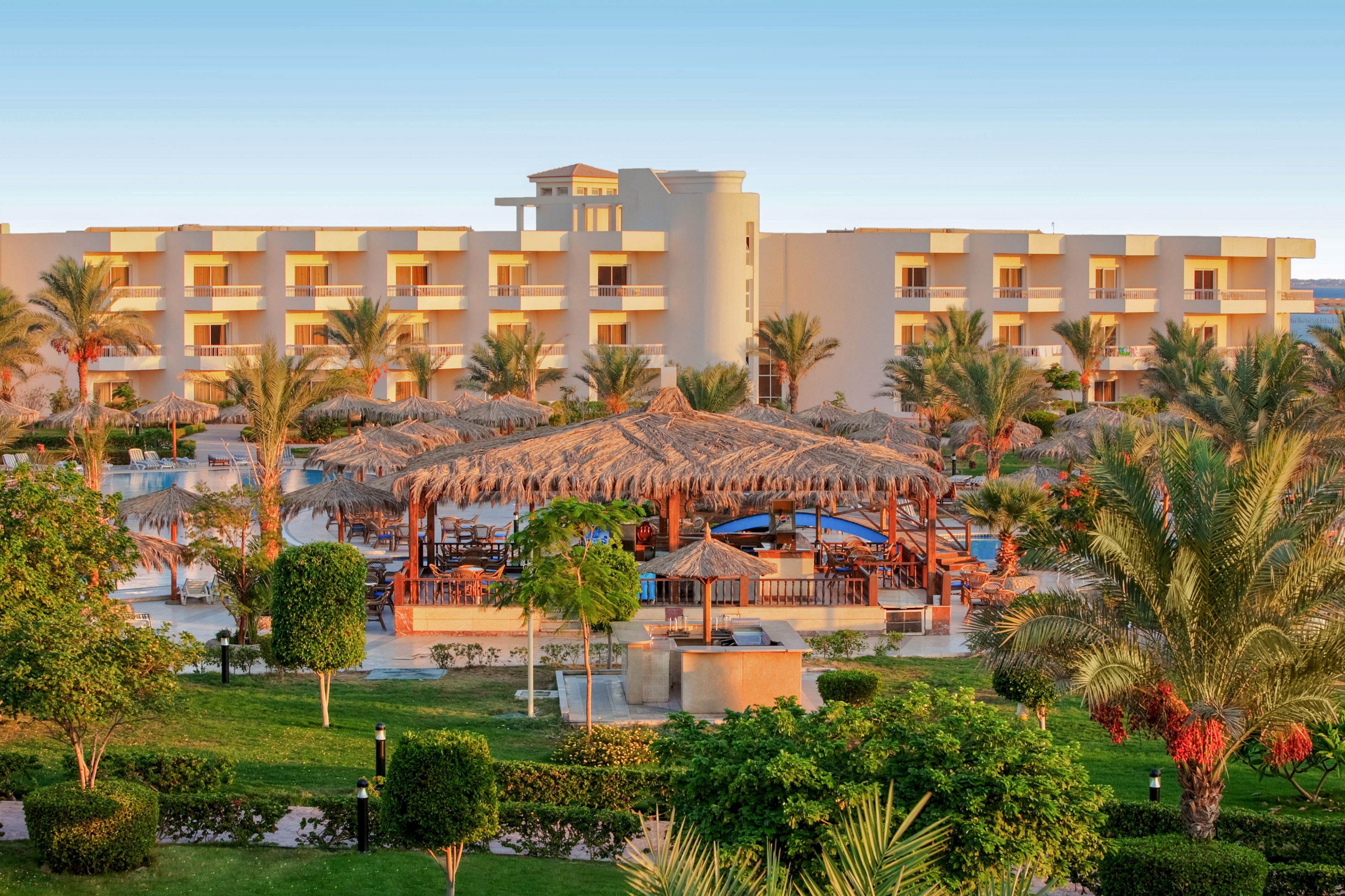 A large resort hotel with multiple balconies overlooks a lush garden and pool area. Thatched-roof cabanas and wooden structures surround the swimming pool, creating a relaxed vacation atmosphere. Palm trees and manicured shrubs fill the foreground, while the building is lit by warm sunset or sunrise light. The scene suggests a tranquil holiday destination in a warm coastal or desert climate.