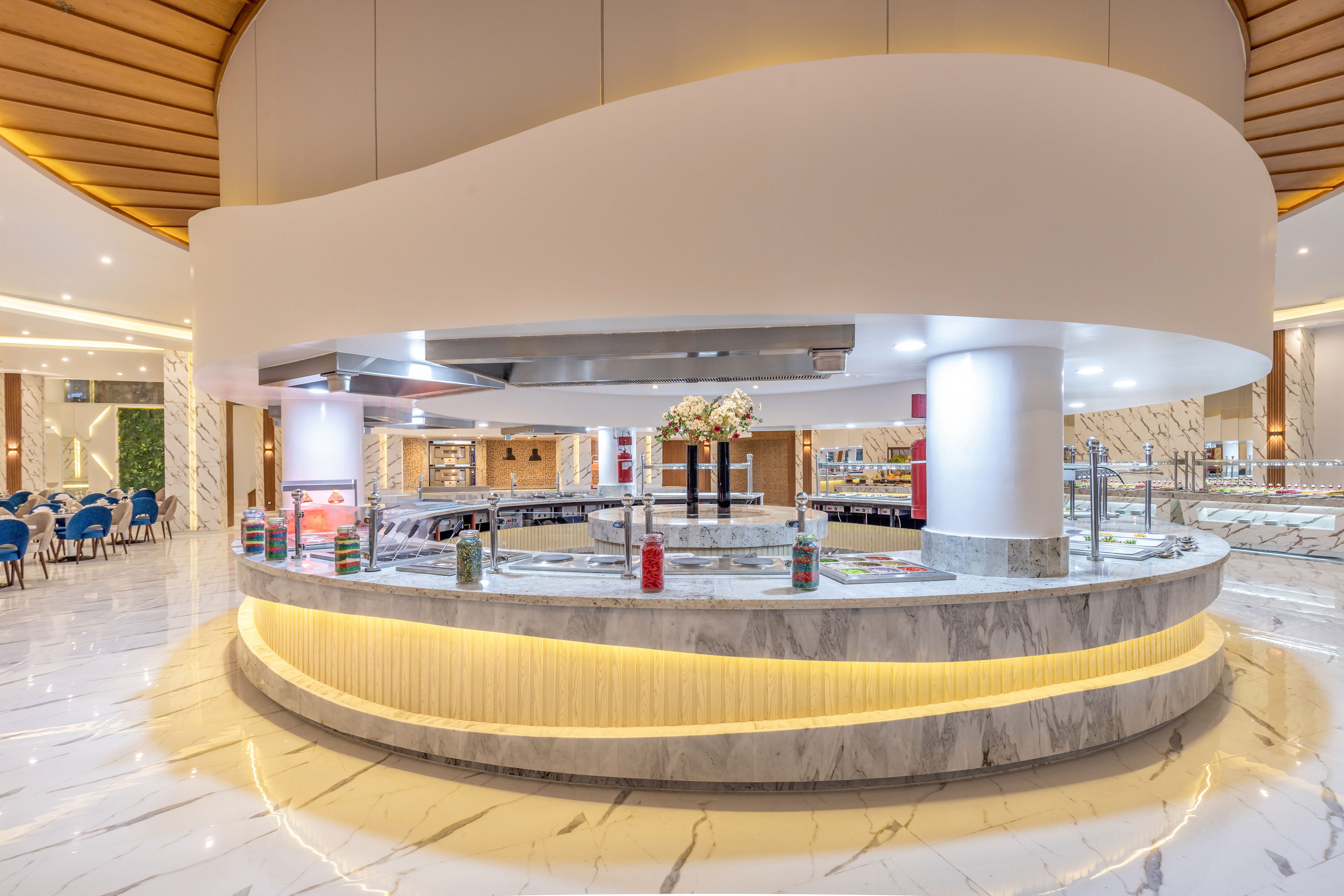 A large circular buffet counter stands in the center of a bright, modern restaurant interior. The counter features a marble finish with warm yellow accent lighting and stainless steel serving stations. Various condiments and bottles are arranged along the edge, with decorative flowers placed on top. The surrounding space includes polished marble floors, contemporary ceiling details, and seating areas with blue chairs.