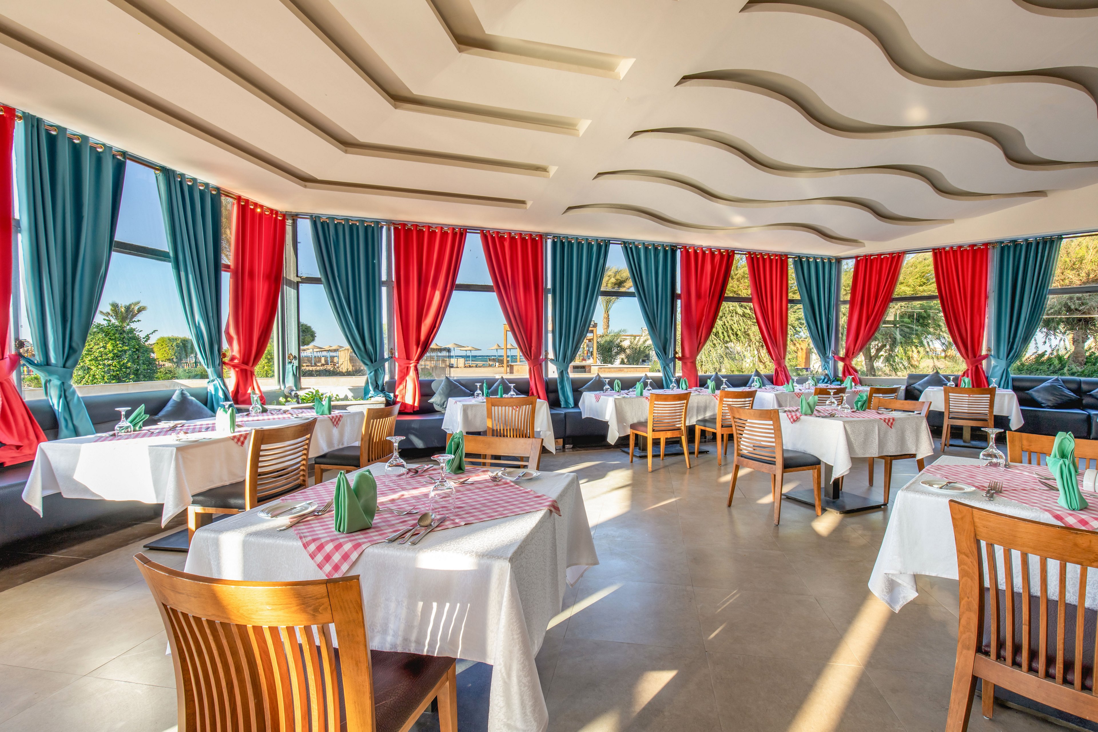A sunlit restaurant dining area features neatly arranged tables with white tablecloths and checkered runners. Wooden chairs surround each table, which is set with glassware, folded napkins, and simple table settings. Large windows reveal an outdoor view with palm trees and greenery, enhancing the airy, coastal atmosphere. Bold teal and red curtains frame the windows, adding vibrant color to the modern interior design.