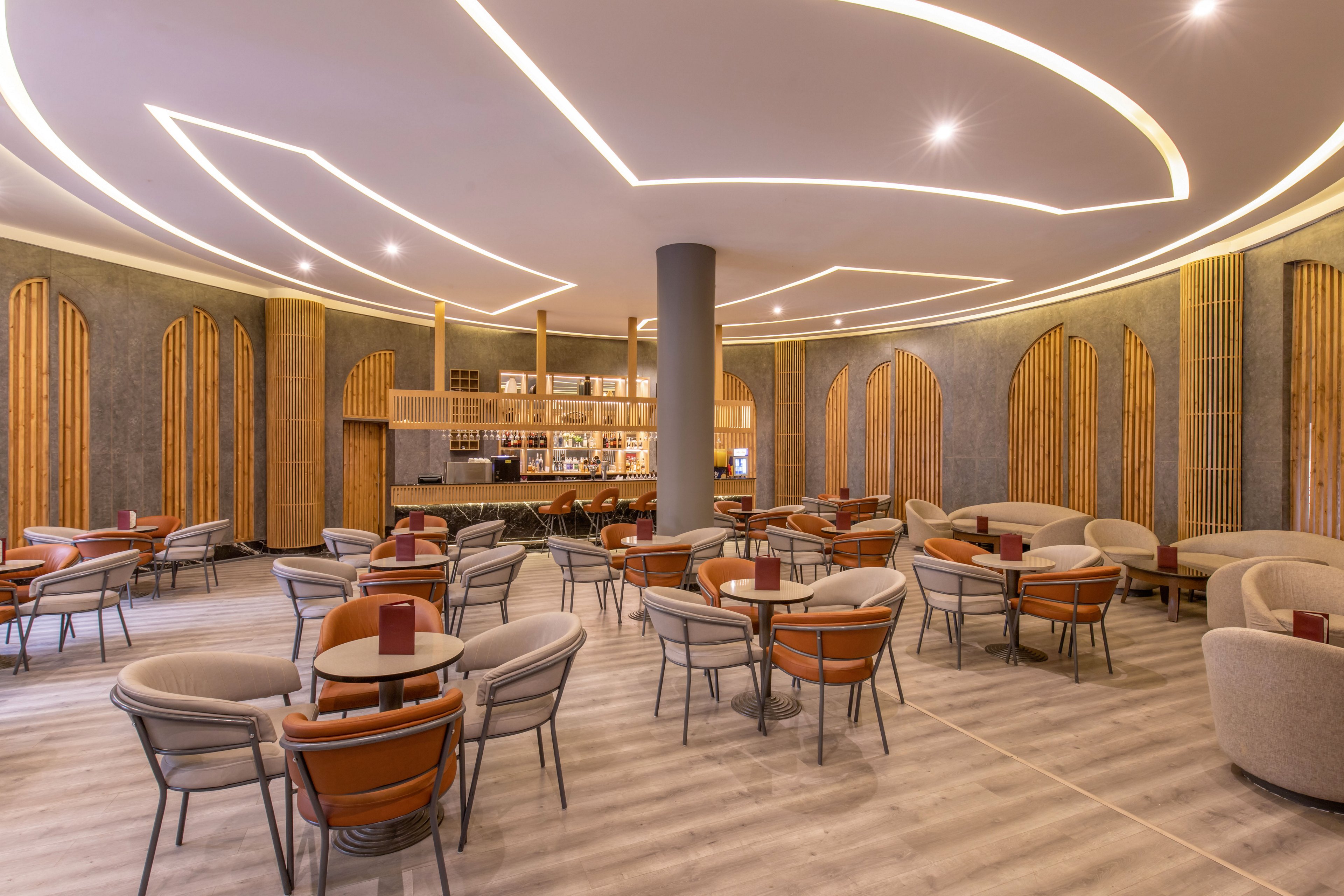 A contemporary lounge bar interior features multiple round tables and cushioned chairs arranged in an open seating plan. Warm wooden wall panels and vertical slats frame the space, complemented by a sleek central column. Curved ceiling light strips and recessed spotlights create a soft, inviting glow across the room. The bar area in the background displays bottles and glassware, while red table menus add subtle color accents.
