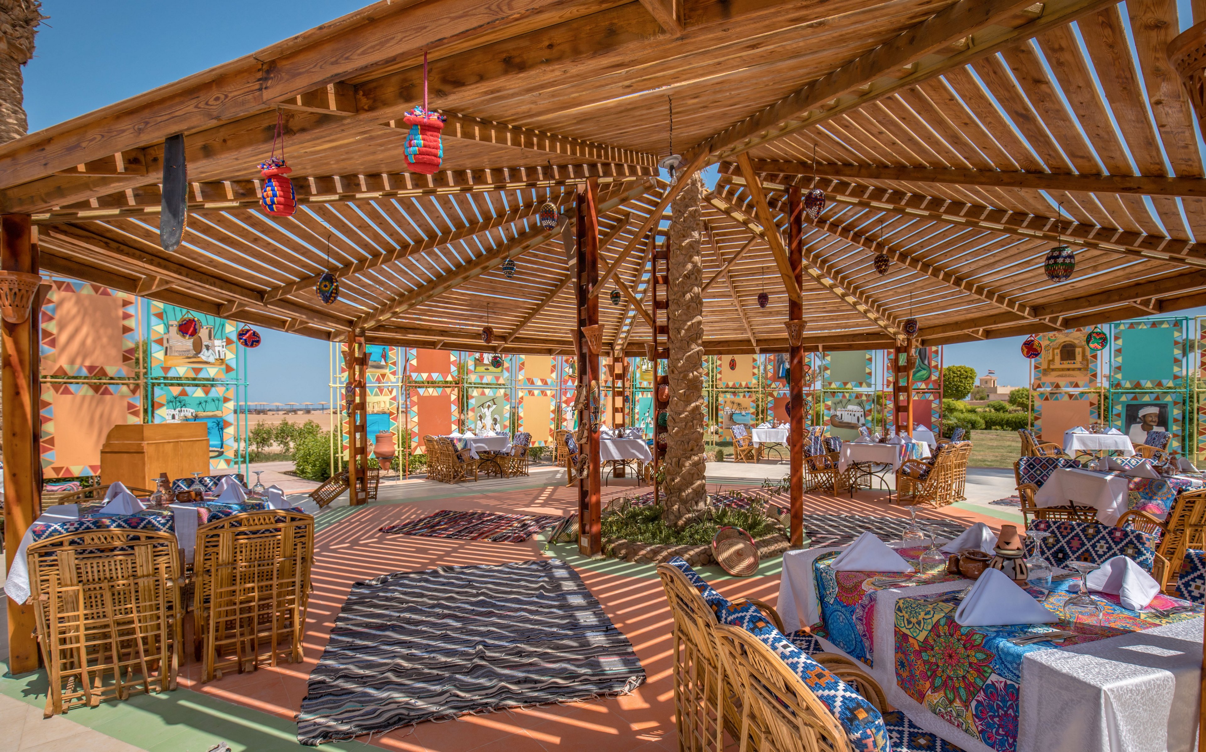 An open-air dining area is set beneath a large wooden pergola with slatted roof casting patterned shadows on the floor. Wicker chairs surround tables covered in white cloths and vibrant, multicolored textiles. Decorative lanterns and hanging ornaments add detail to the rustic wooden structure. Brightly painted walls and clear blue sky in the background create a warm, inviting atmosphere.
