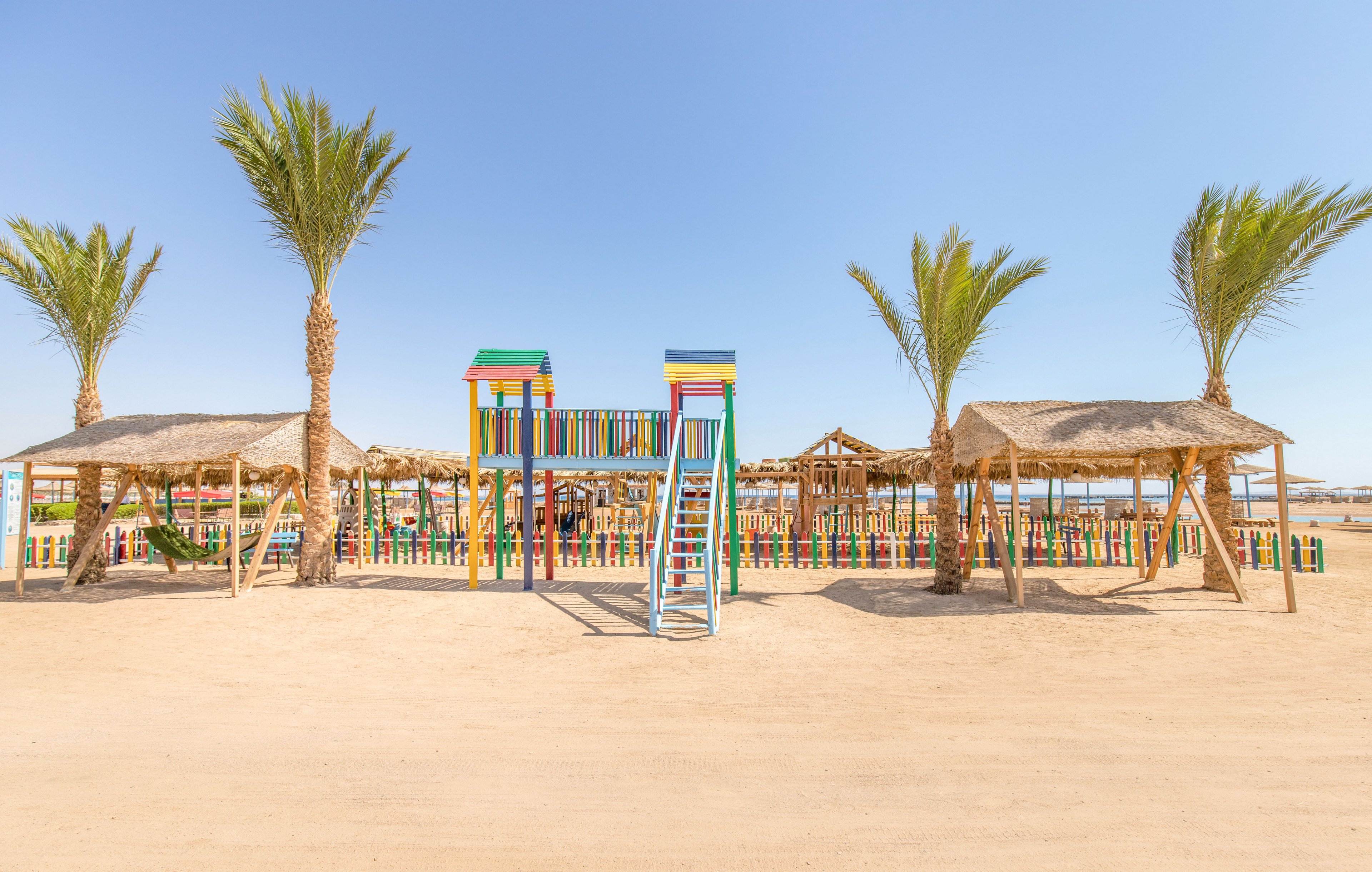 A vibrant playground with multicolored structures stands on a sandy beach under a clear blue sky. Palm trees and thatched shade huts frame the play area, creating a tropical seaside setting. The playground features a slide, elevated platforms, and vertical bars painted in bright colors. In the background, calm water and additional wooden structures suggest a family-friendly coastal resort.