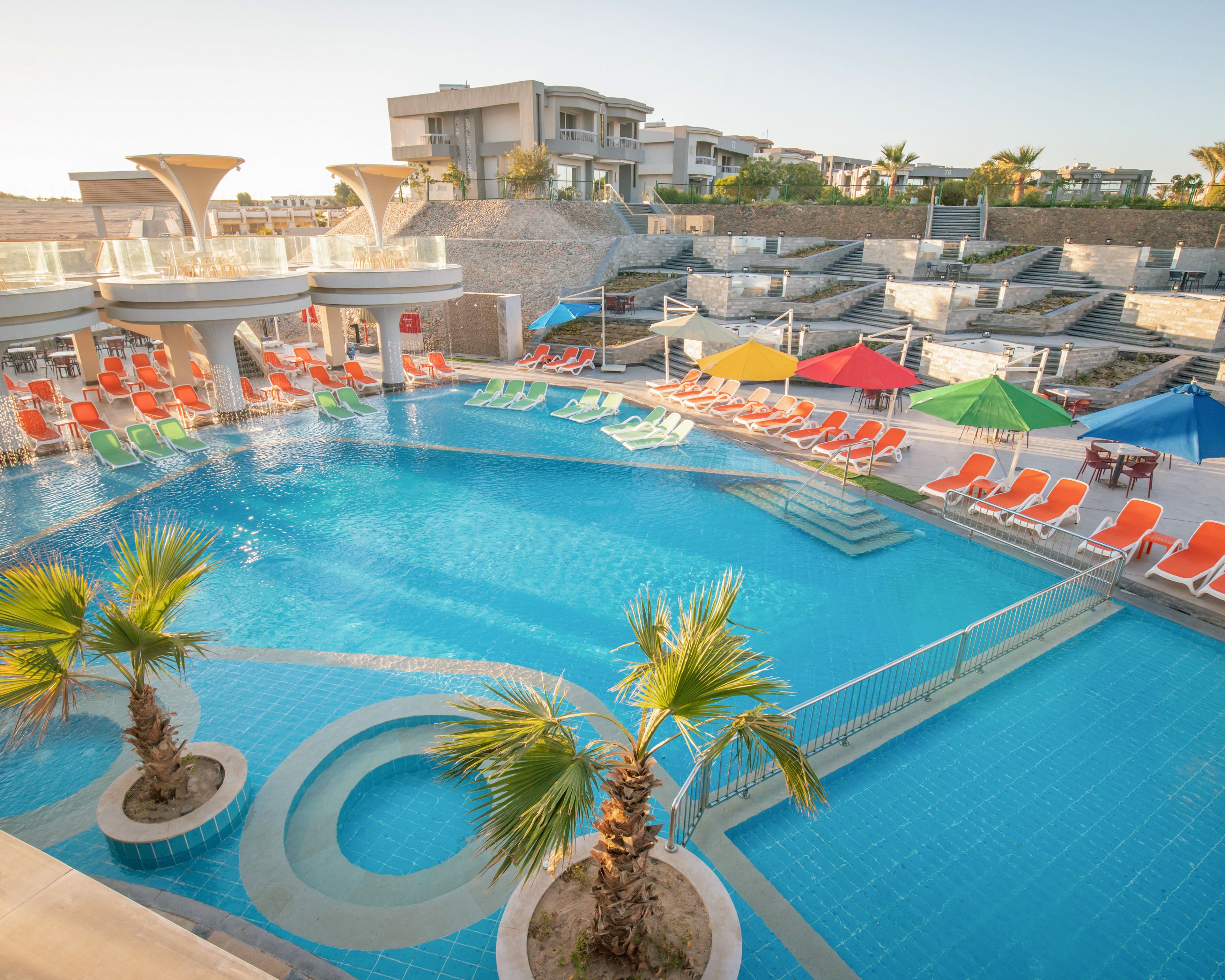 A large resort swimming pool is surrounded by rows of bright orange, green, and yellow loungers. Colorful umbrellas line the poolside, creating a vibrant and inviting atmosphere. Two small palm trees stand on decorative islands within the clear blue water. Modern multi-story buildings and terraces rise in the background under a clear sky, suggesting a warm vacation destination.