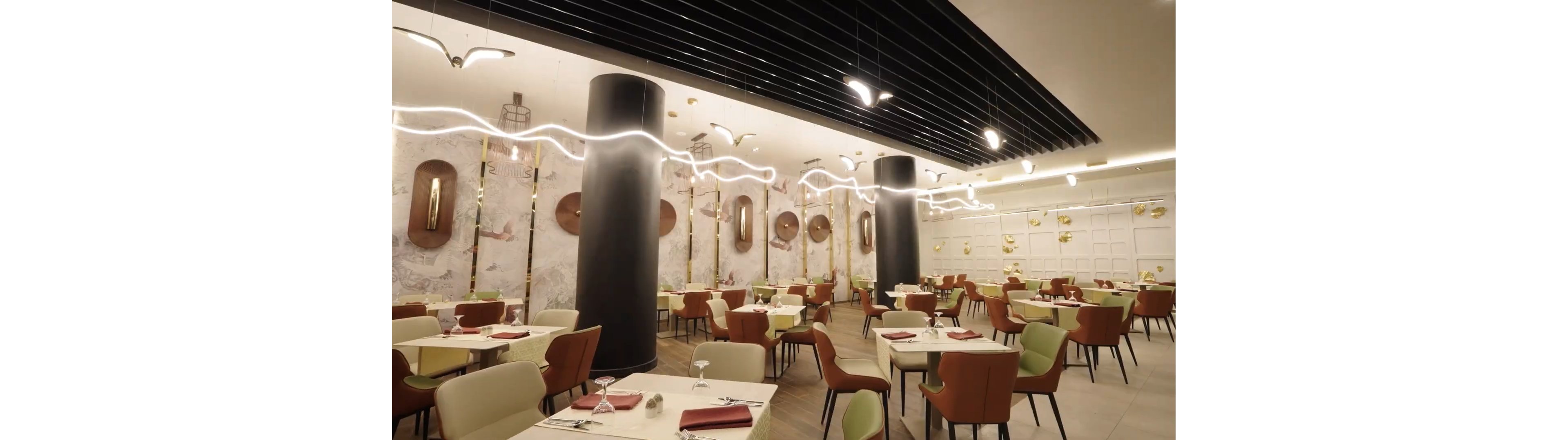A contemporary restaurant dining room features neatly arranged tables with beige and light green chairs. Each table is set with glassware, napkins, and simple place settings, ready for guests. Large black columns and a wavy ceiling light installation create a striking visual focal point. Warm wall decor and soft lighting contribute to an elegant, inviting atmosphere.