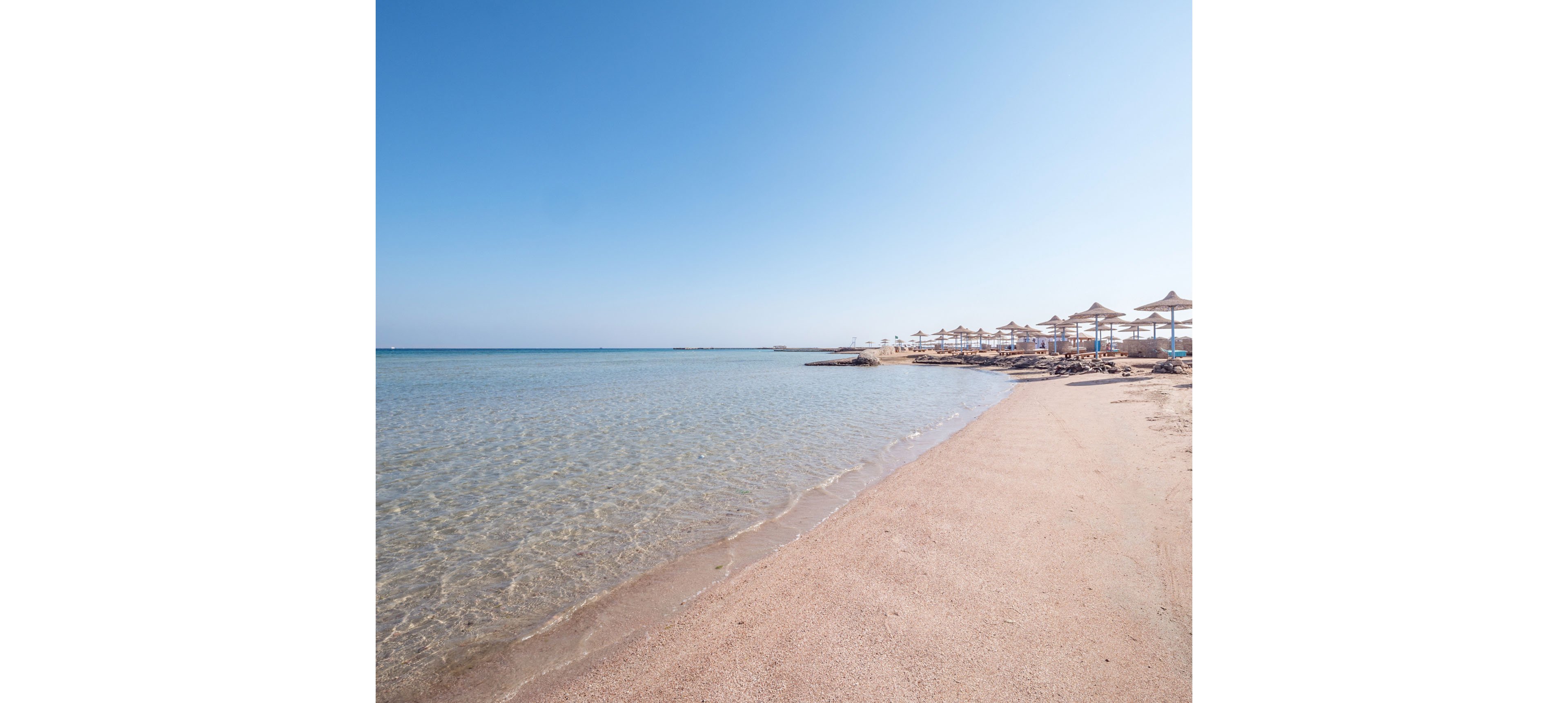 A tranquil sandy beach stretches along clear, shallow turquoise water under a bright blue sky. Straw sun umbrellas line the shore in the distance, suggesting a quiet resort or holiday destination. The scene is calm and minimal, with no people visible and soft waves gently meeting the sand. The wide horizon and open space create a peaceful, relaxing atmosphere.