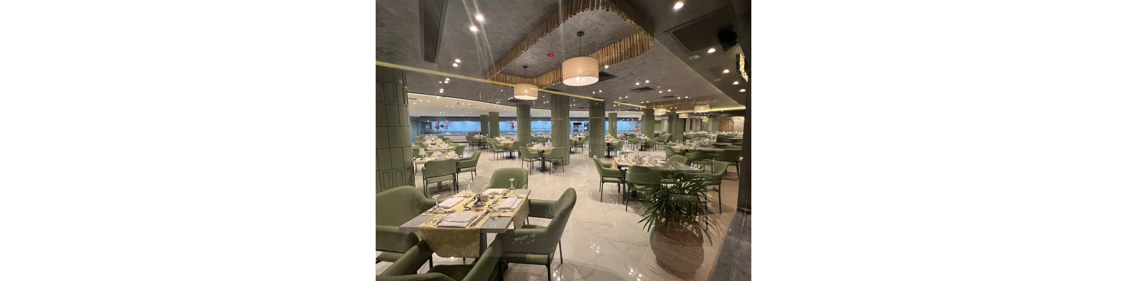 Spacious restaurant interior features neatly arranged tables with green upholstered chairs and polished place settings. The room has a contemporary design with large columns, hanging pendant lights, and reflective ceiling details. Bright lighting and glossy tiled floors create an airy, upscale atmosphere. Tables are set for service, suggesting a fine dining experience in a modern urban setting.
