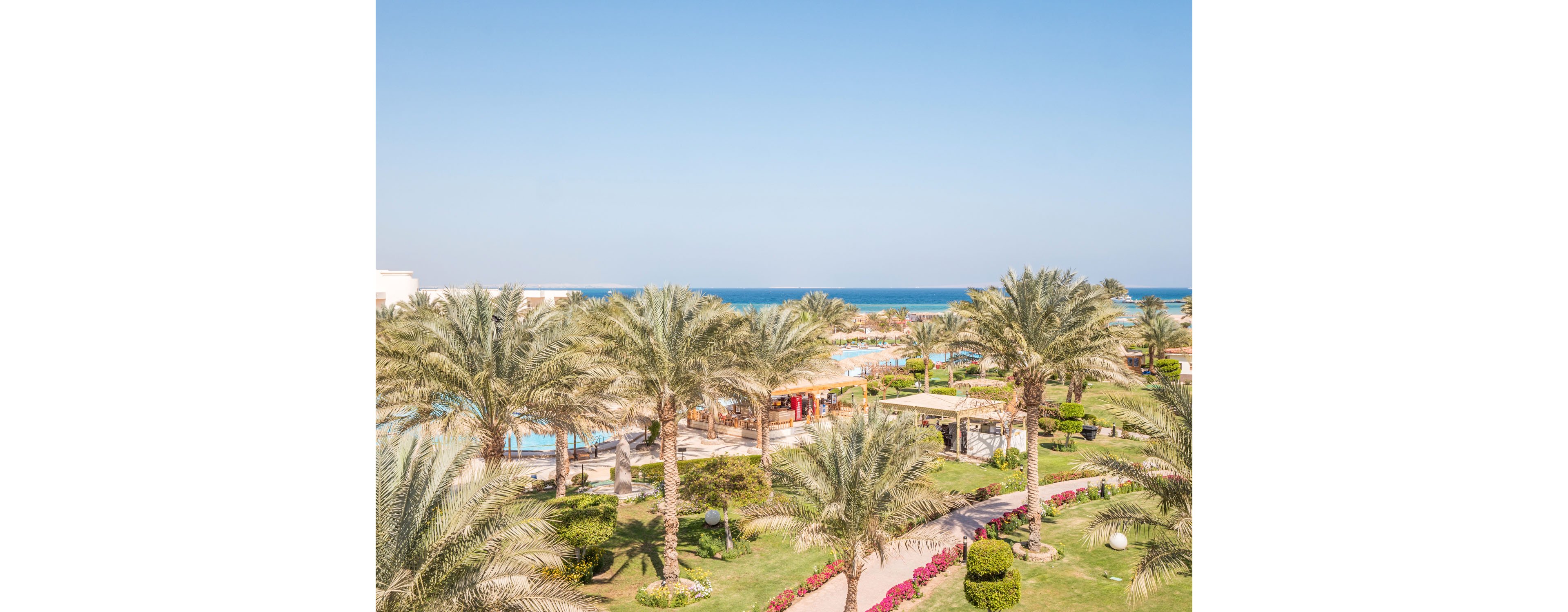 A bright coastal resort landscape features tall palm trees and manicured green lawns. Walking paths curve through gardens with colorful flowers and trimmed bushes. Several swimming pools and sun loungers are visible among low buildings and shaded areas. In the background, the calm blue sea meets a clear sky, creating a relaxed vacation atmosphere.