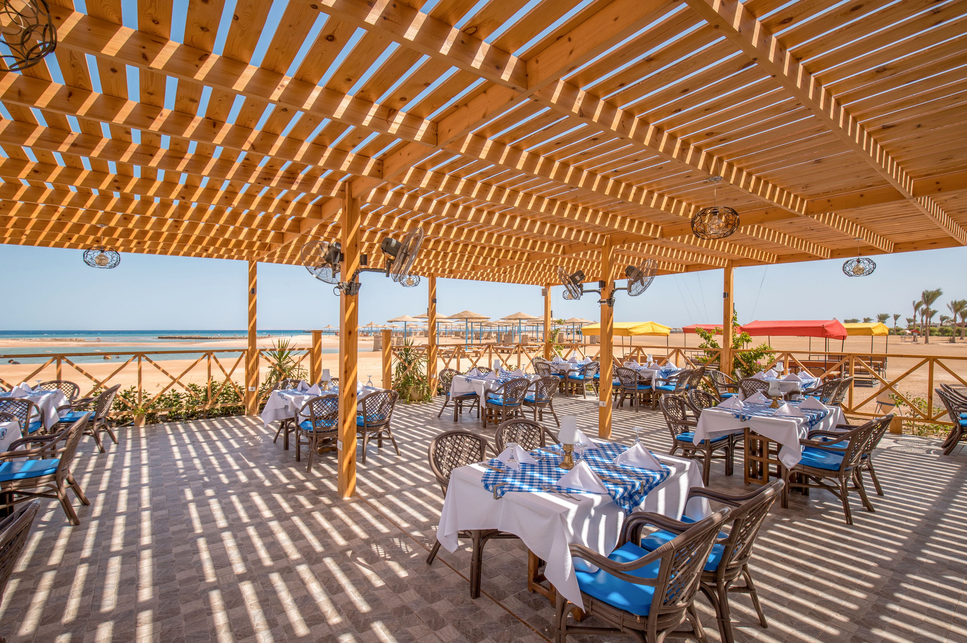 An open-air seaside restaurant is set beneath a large wooden pergola casting striped shadows across the tiled floor. Tables are neatly arranged with white tablecloths and blue checkered accents, facing the sandy beach and ocean. Wicker-style chairs surround each table, and decorative lanterns hang from the pergola beams. Colorful beach huts and palm trees are visible in the background, enhancing the relaxed coastal atmosphere.