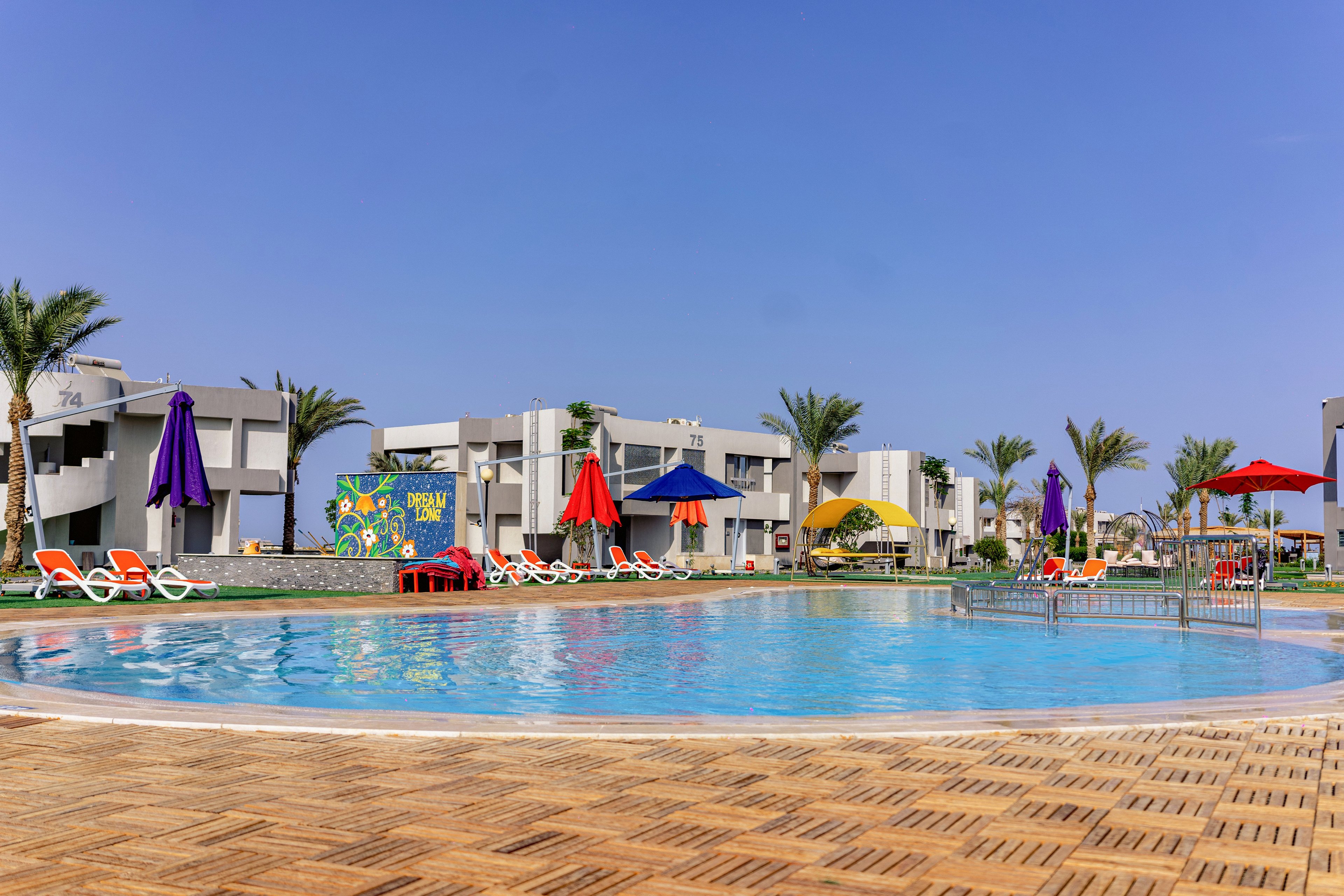A large circular swimming pool sits at the center of a modern resort complex. Colorful sun umbrellas and lounge chairs surround the pool on a wooden deck, creating a relaxed vacation atmosphere. White low-rise buildings and palm trees frame the scene under a clear blue sky. The area appears calm and inviting, suggesting a sunny holiday destination.