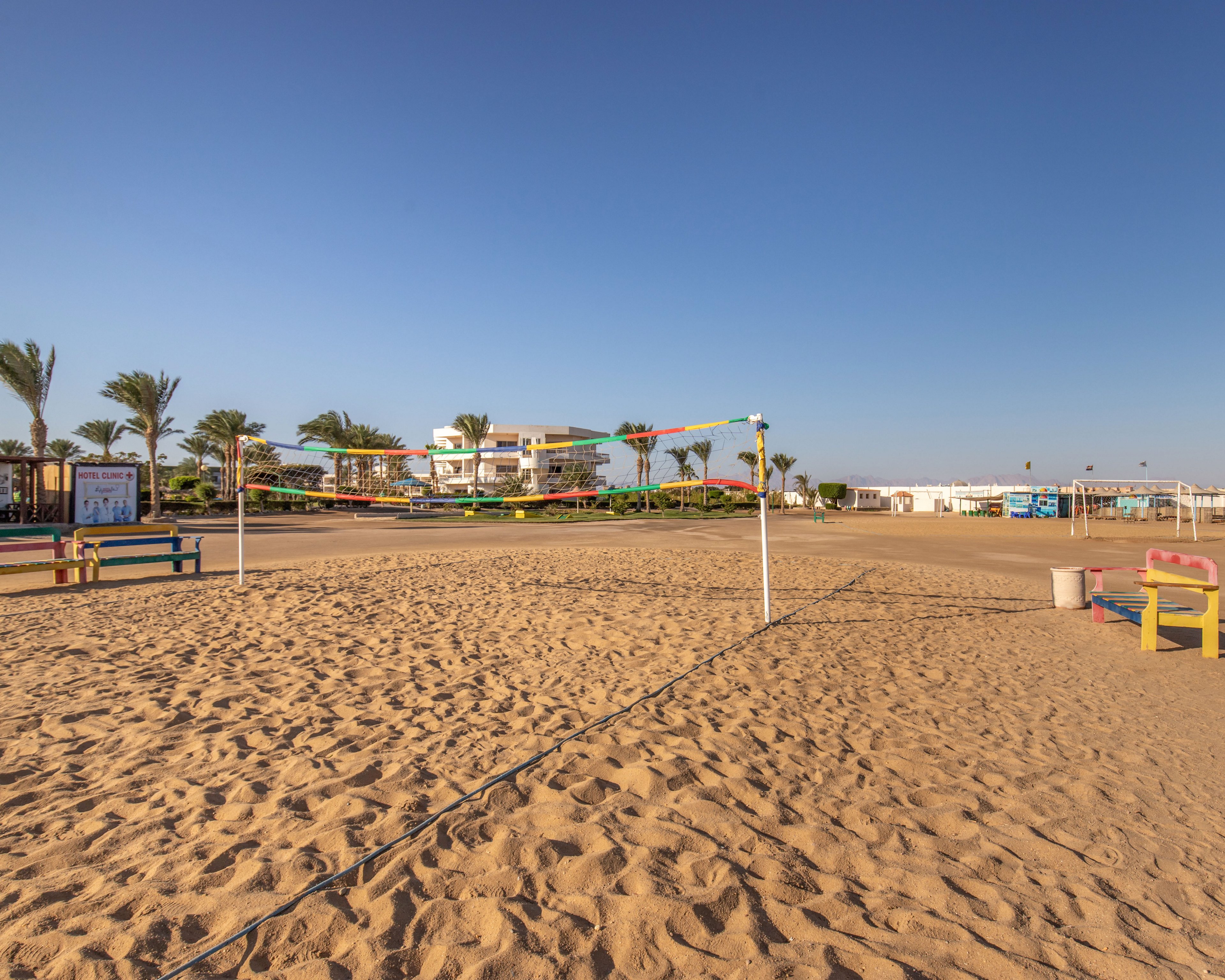 A sandy beach volleyball court stands empty under a clear blue sky at a seaside resort. Colorful nets and boundary lines define the play area, with benches and small structures visible around the court. Palm trees and low-rise resort buildings appear in the background, suggesting a relaxed holiday destination. The warm light and long shadows indicate early morning or late afternoon on a calm day.