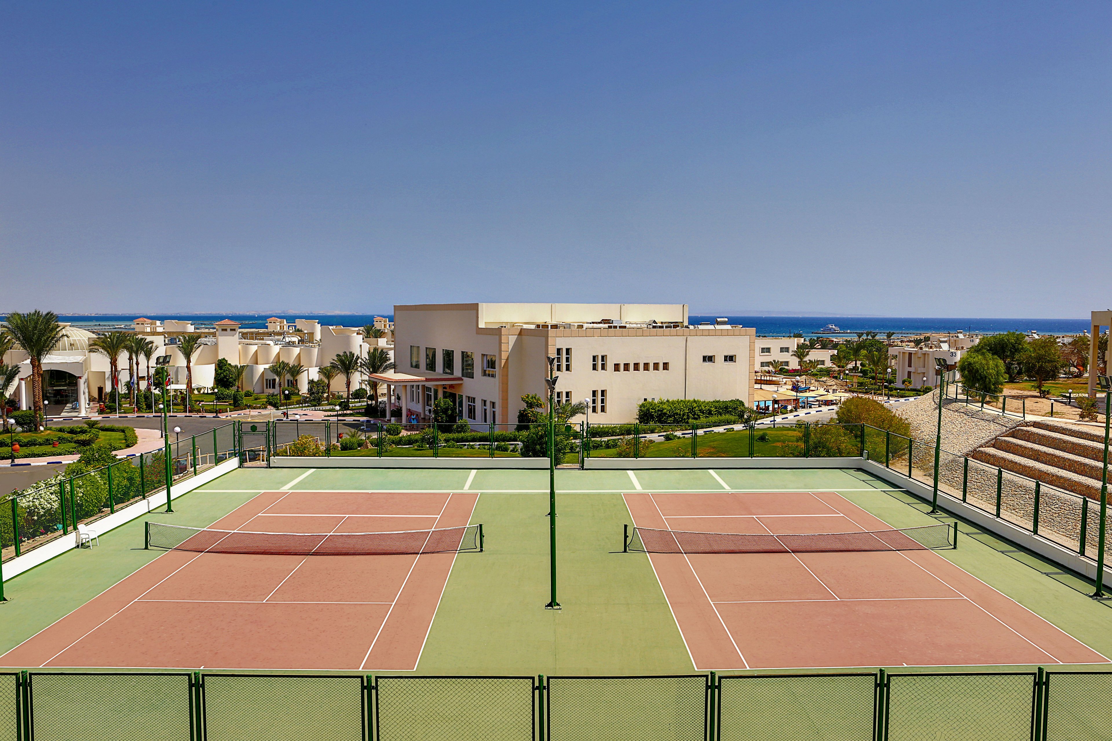 Two empty tennis courts sit in the foreground of a sunny resort complex. Low-rise white buildings and landscaped paths surround the sports area, leading toward a distant coastal town. The blue sea and clear sky create a bright, open backdrop. The scene suggests a vacation destination with outdoor recreation facilities and scenic ocean views.