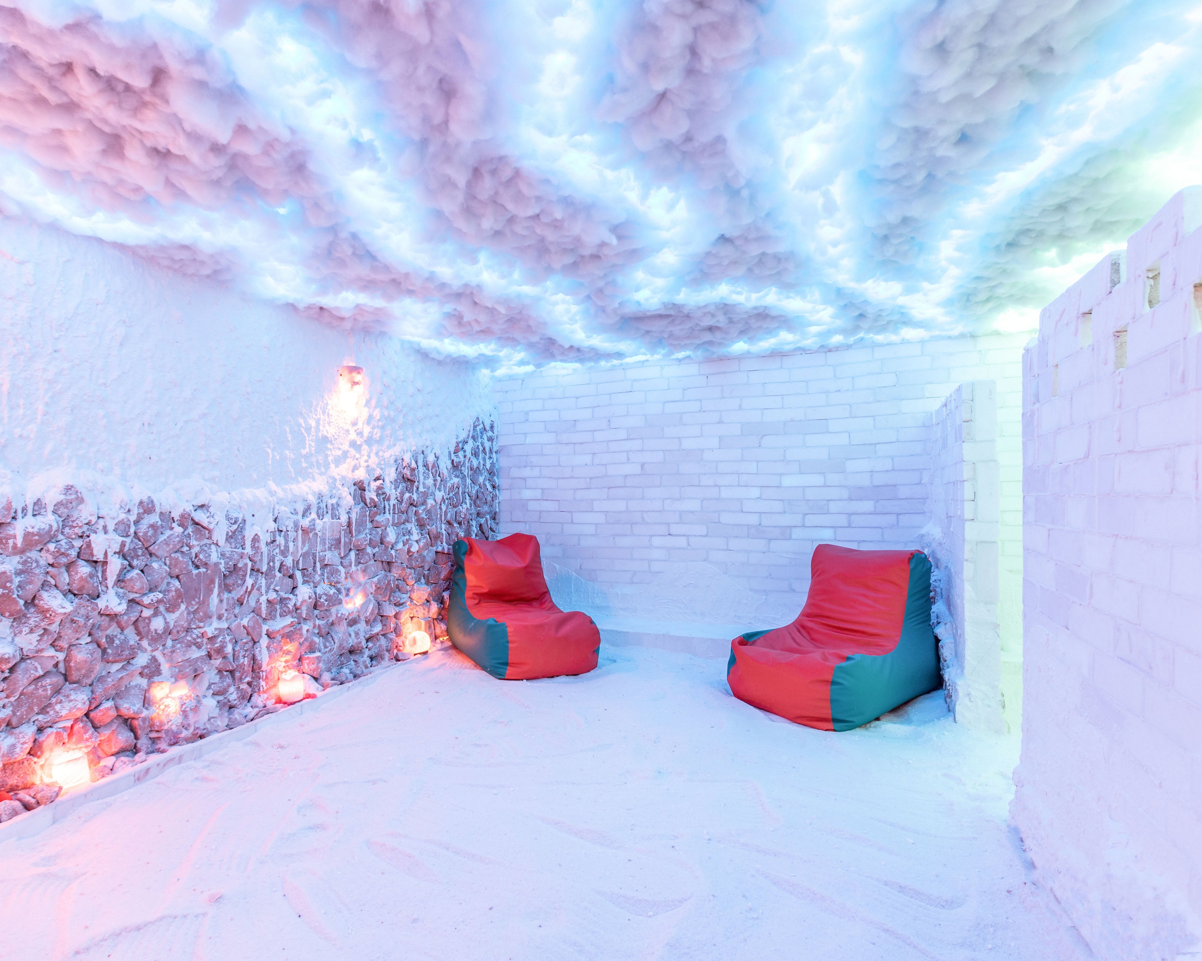 A bright, contemporary ice room features two colorful lounge chairs set against white brick and stone walls. The floor and ceiling are covered in snow-like material, creating a winter cave atmosphere. Soft colored lighting glows from fixtures along the rock wall and above, casting blue and pink hues throughout the space. The scene feels cozy and surreal, blending cold textures with warm, inviting seating.