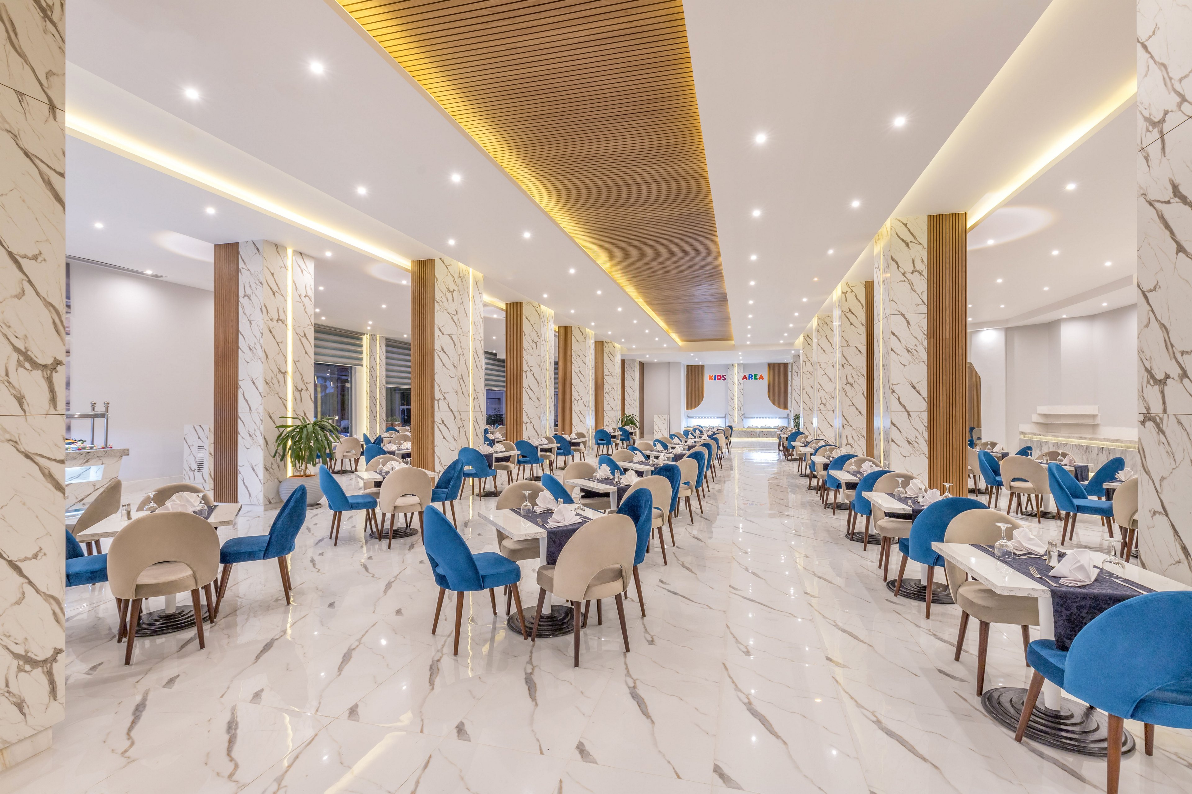 A spacious contemporary restaurant dining area features rows of tables and blue upholstered chairs. The room is lined with tall marble-clad columns and a glossy marble floor that reflects the ceiling lights. Warm wooden ceiling panels and neutral walls create a clean, elegant atmosphere. Tables are neatly set with white linens and tableware, suggesting readiness for guests in a refined setting.