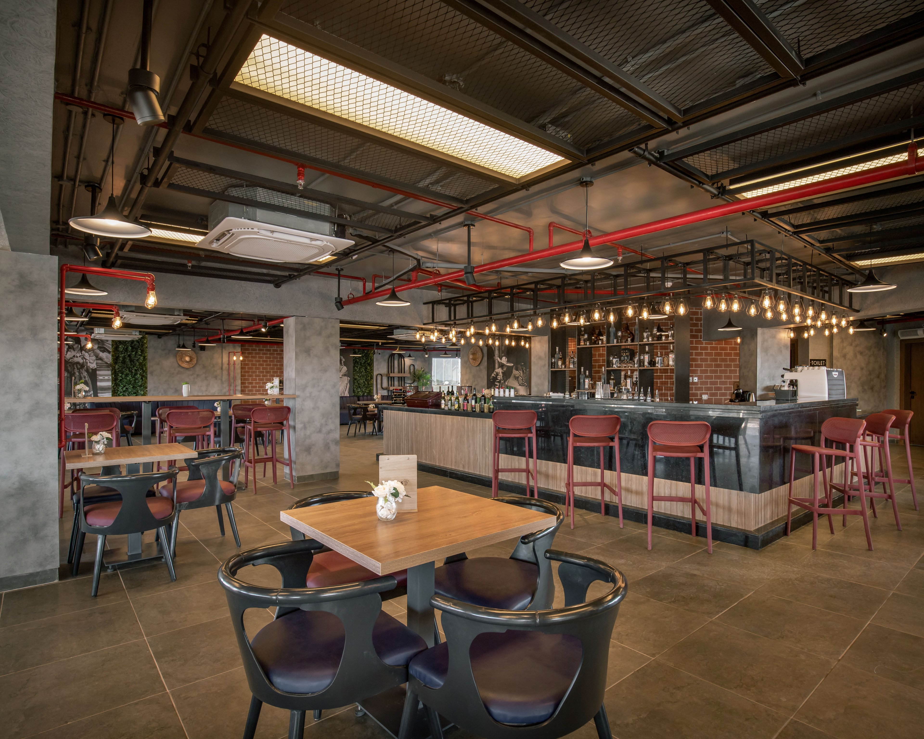 A contemporary bar interior features a central counter surrounded by high stools and tables. The space has an industrial design with exposed ceiling pipes, concrete columns, and warm pendant lighting above the bar. Several dining tables and chairs are arranged across the tiled floor, with a small flower arrangement on the nearest table. The atmosphere appears relaxed and inviting, suitable for socializing or casual dining.
