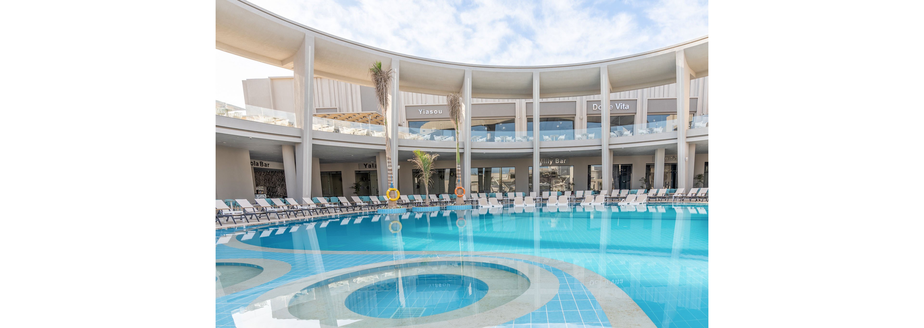 A large circular swimming pool sits at the center of a modern resort courtyard. White lounge chairs line the curved poolside deck beneath an open, colonnaded terrace. Bar and restaurant signs such as "Yali Bar" and "Dolce Vita" are visible along the upper level. The scene is bright and tranquil under a partly cloudy sky, emphasizing leisure and vacation relaxation.