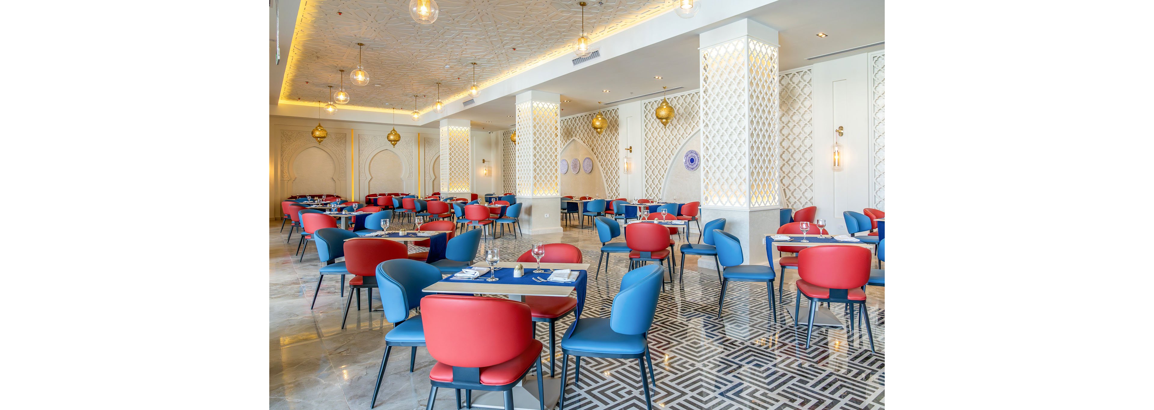 A spacious, contemporary restaurant dining area features rows of tables with red and blue upholstered chairs. The setting includes decorative white lattice columns, patterned floor tiles, and warm pendant lighting hanging from the ceiling. Gold-toned wall sconces and subtle arch motifs add an elegant, slightly luxurious atmosphere. The room appears ready for guests, with neatly arranged place settings on each table.