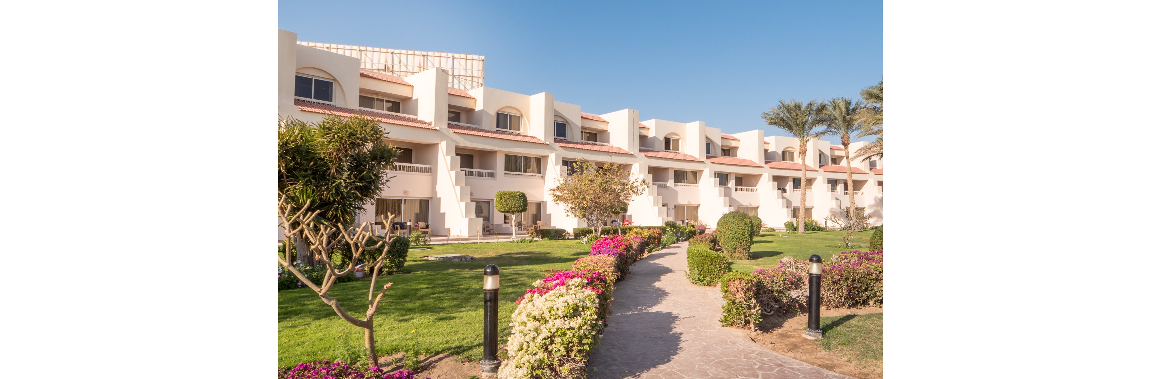 A row of modern, cream-colored resort apartments stands under a clear blue sky. A stone walkway leads through a lush, manicured garden filled with green lawns, flowering shrubs, and trimmed trees. Black bollard lights line the path, adding structure and guiding visitors toward the buildings. The overall scene feels bright, peaceful, and inviting, suggesting a warm vacation destination.