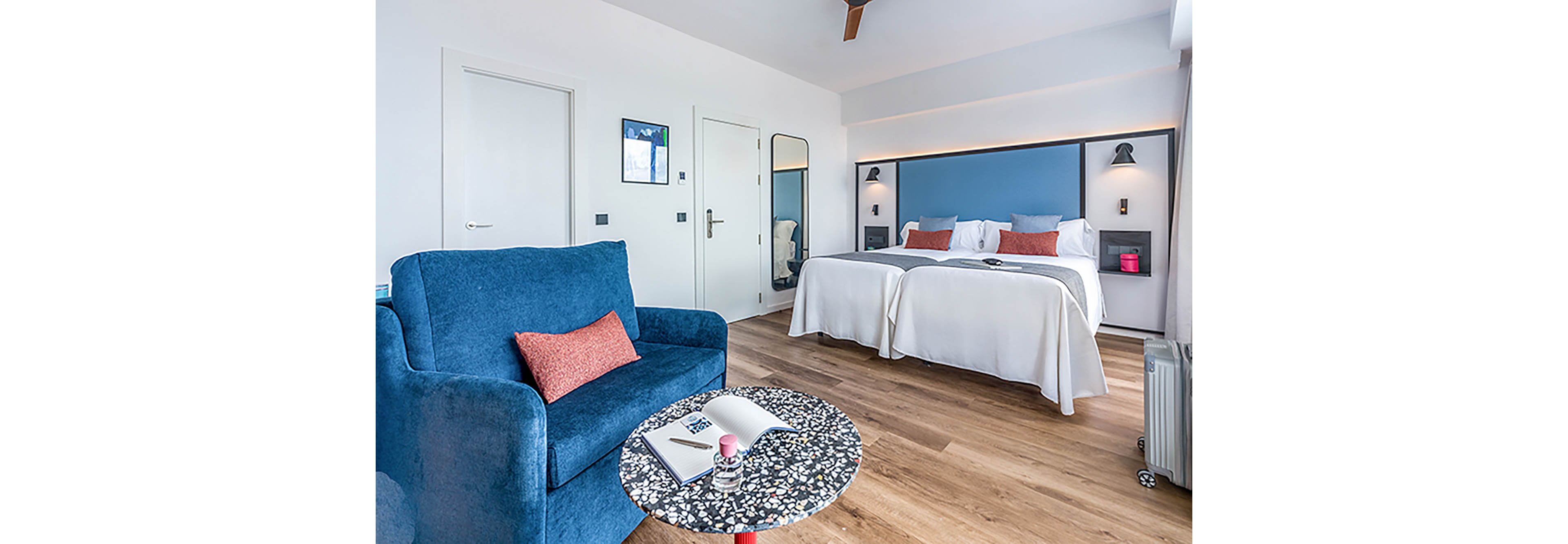 A contemporary hotel room features two neatly made twin beds against a blue accent wall. The space includes a blue armchair with a red cushion and a small round table holding an open notebook and cup. Wooden flooring and white walls create a bright, minimalist atmosphere. Soft lighting and simple decor contribute to a clean, comfortable, and inviting interior.